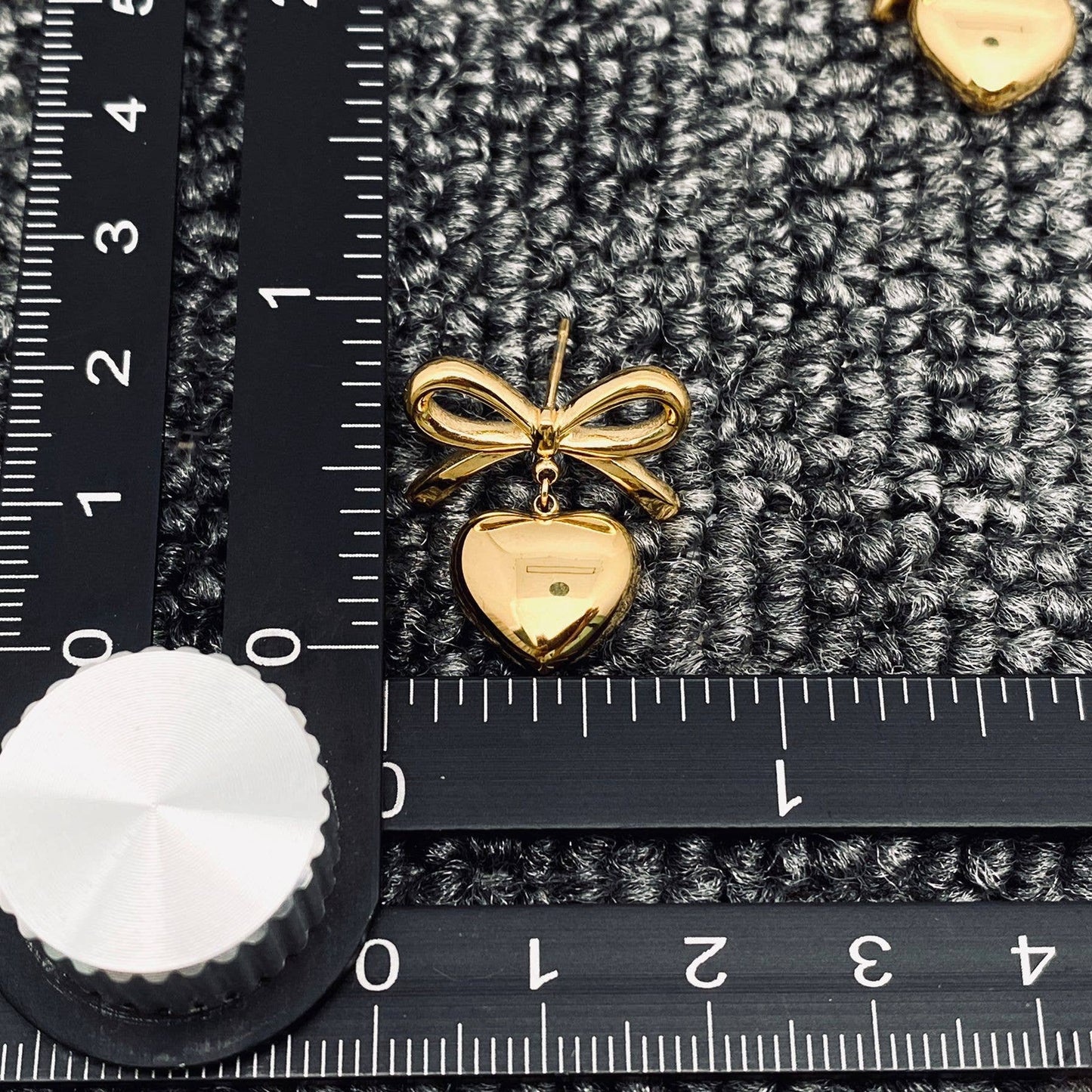 a single gold bow and heart dangle earring next to a black ruler laying on grey carpet
