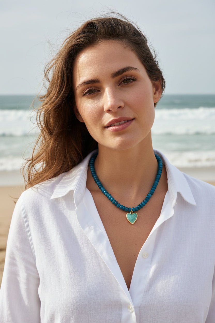 A woman wearing a bllue beaded necklace with a heart-shaped pendant in a beach setting.