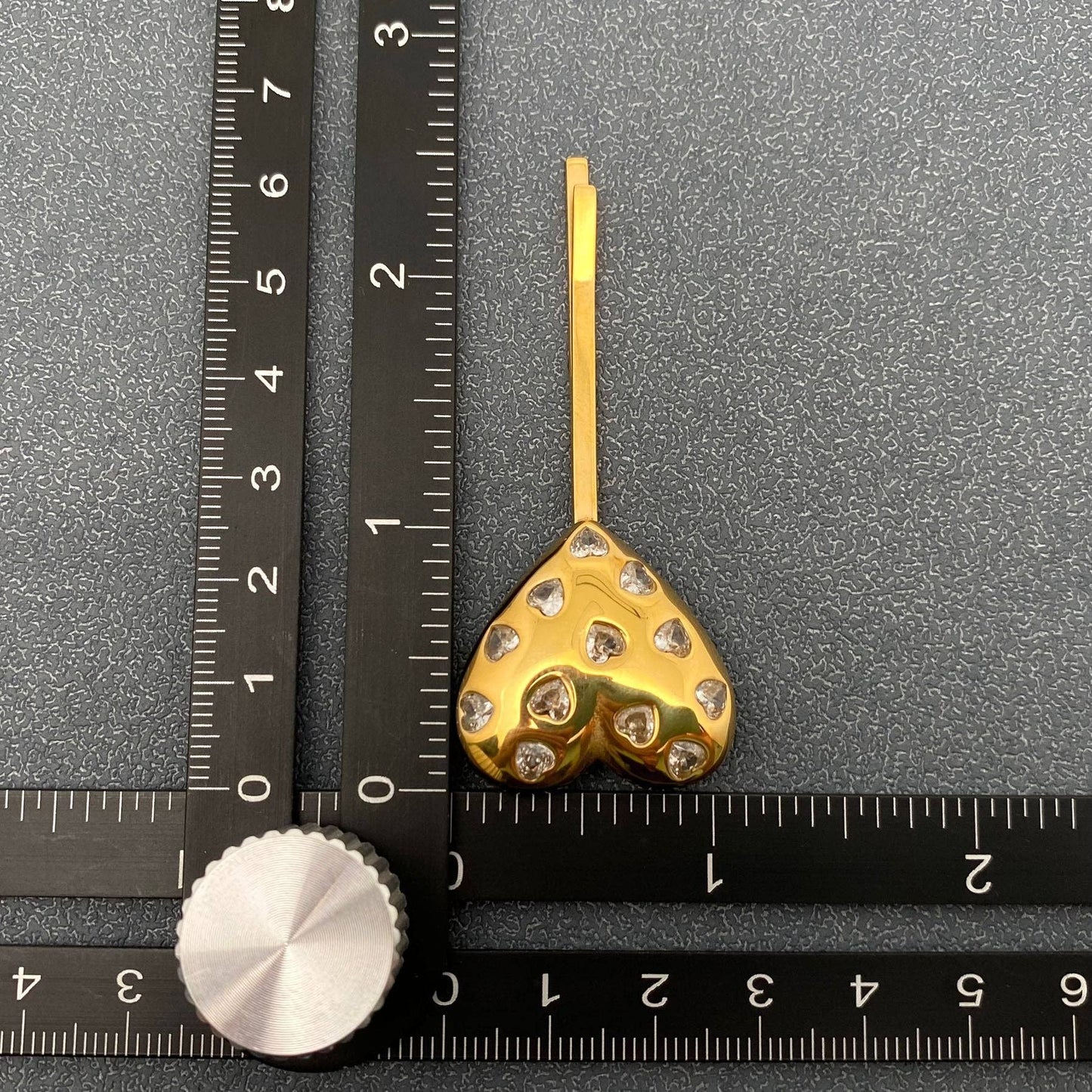 A gold heart hair pin with inlaid rhinestones next to a rule for scale on a grey background