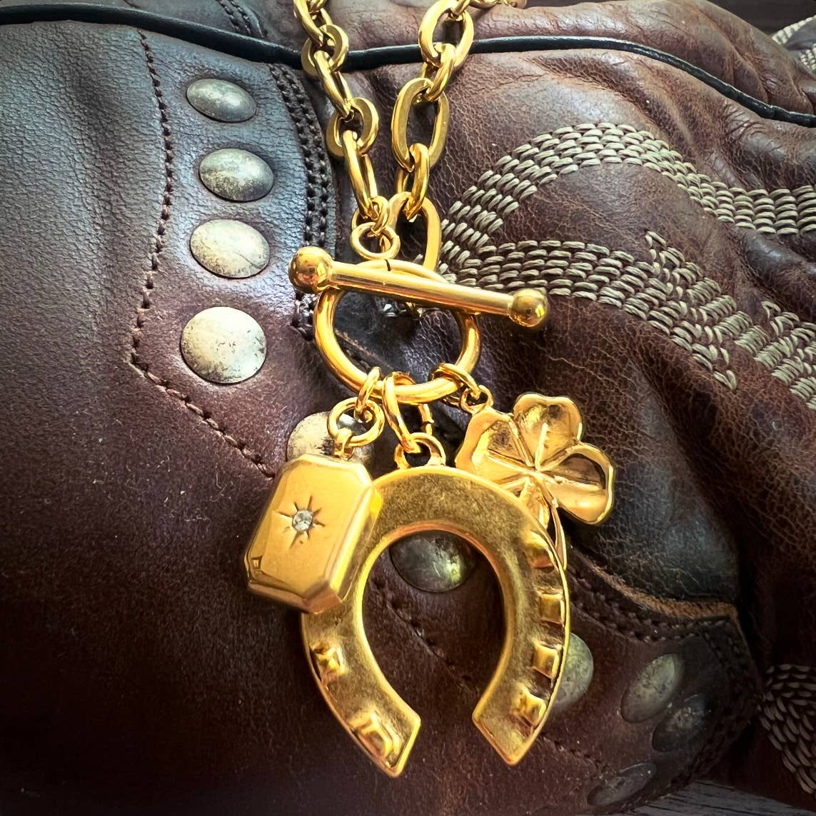 Gold multi charm chain necklace with horseshoe, clover, and rectangle pendant laying an a western style brown boot