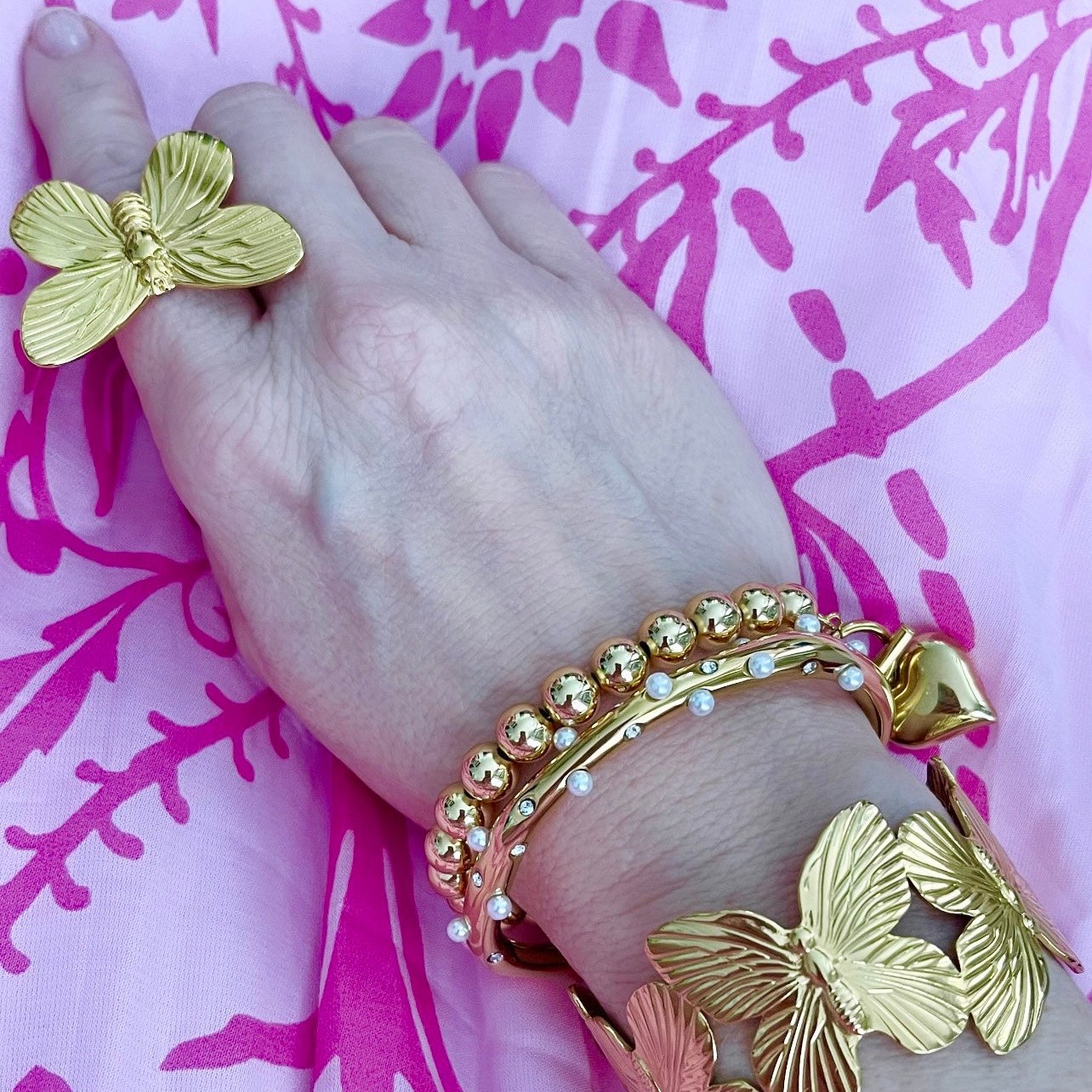 woman in pink dress wearing gold bead bracelet with puffy heart charm and toggle clasp, gold butterly ring, a pearl encrusted gold bracelet, and a gold butterfly cuff