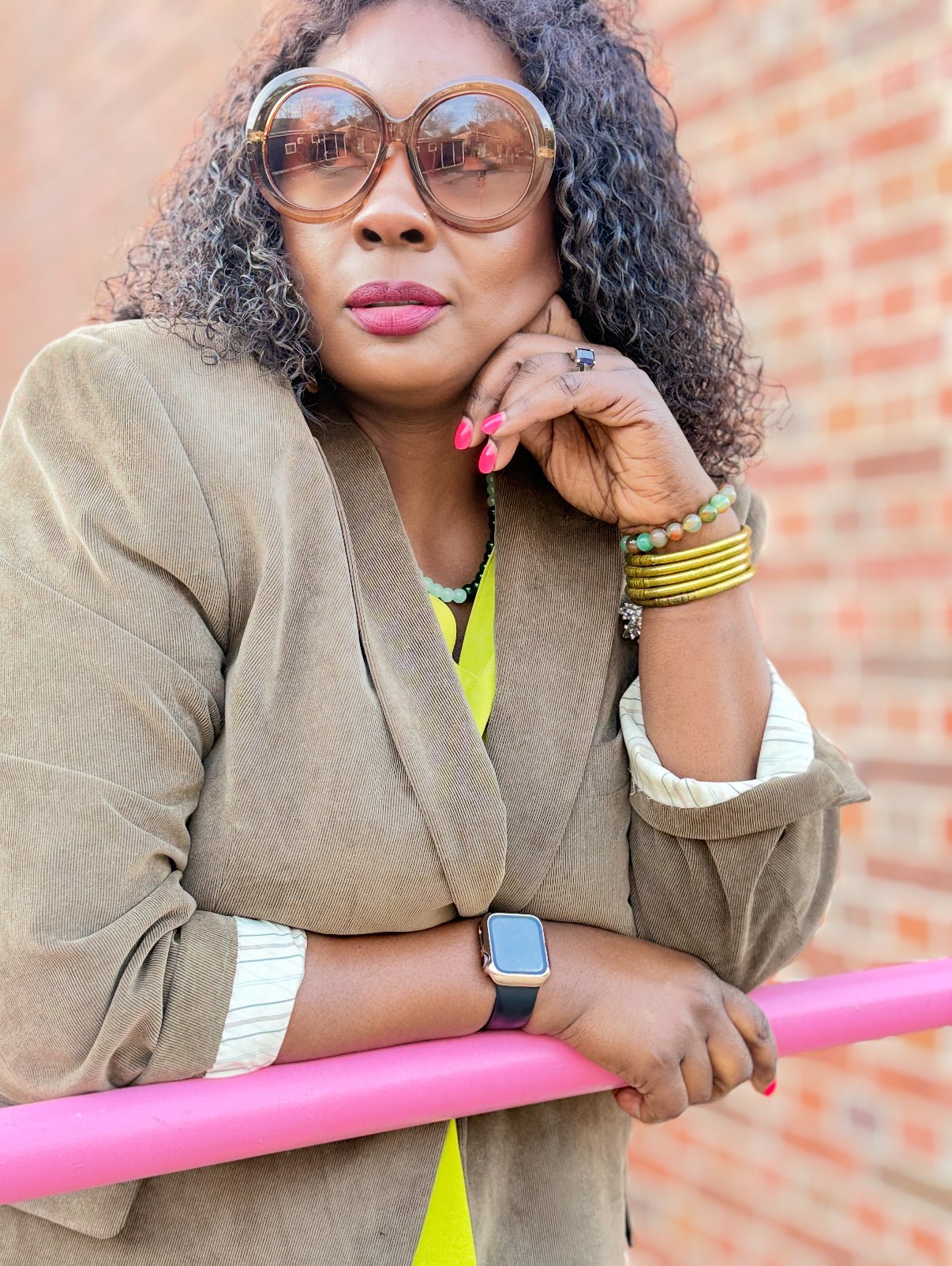 Woman wearing sunglasses and a beige jacket leaning on a pink hand railing outside with a brick wall background