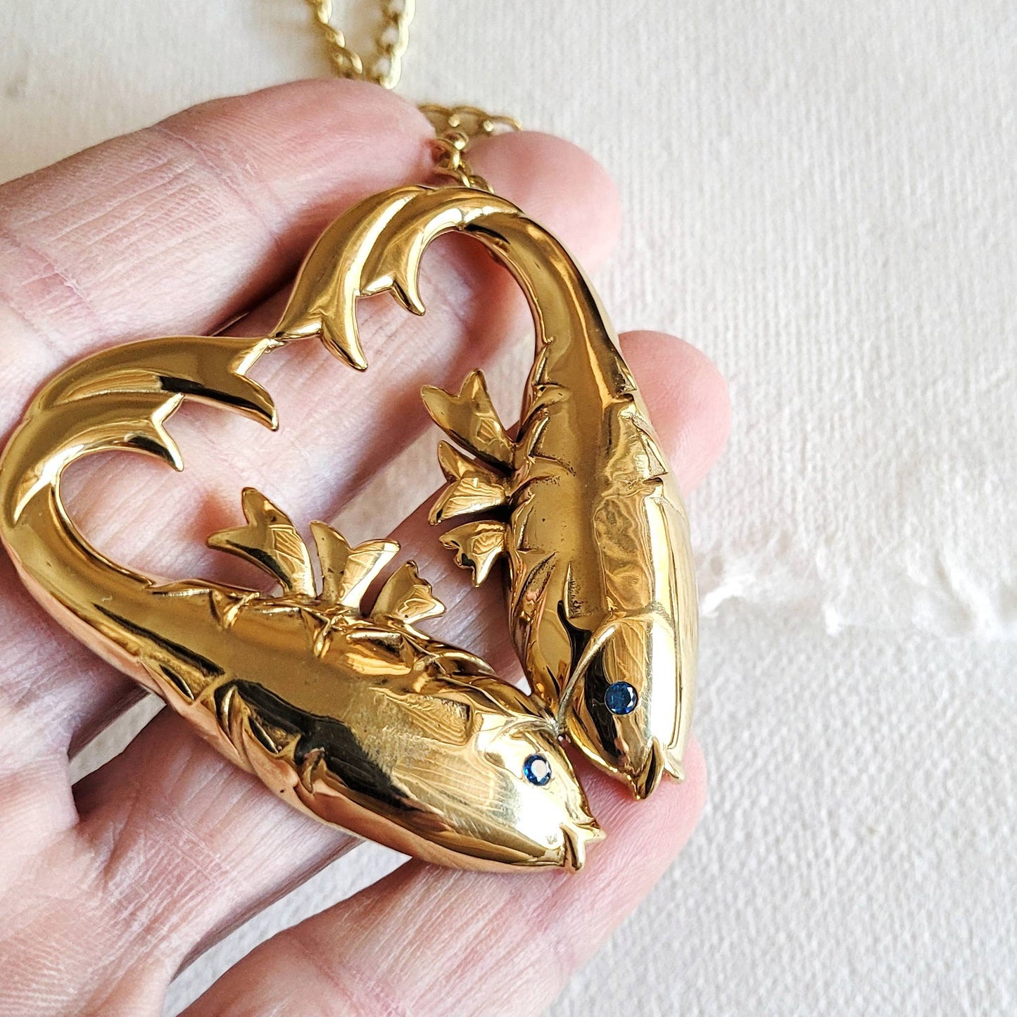 gold double fish heart shaed necklace with aquamarine inlaid stones for the eyes in the palm of a hand