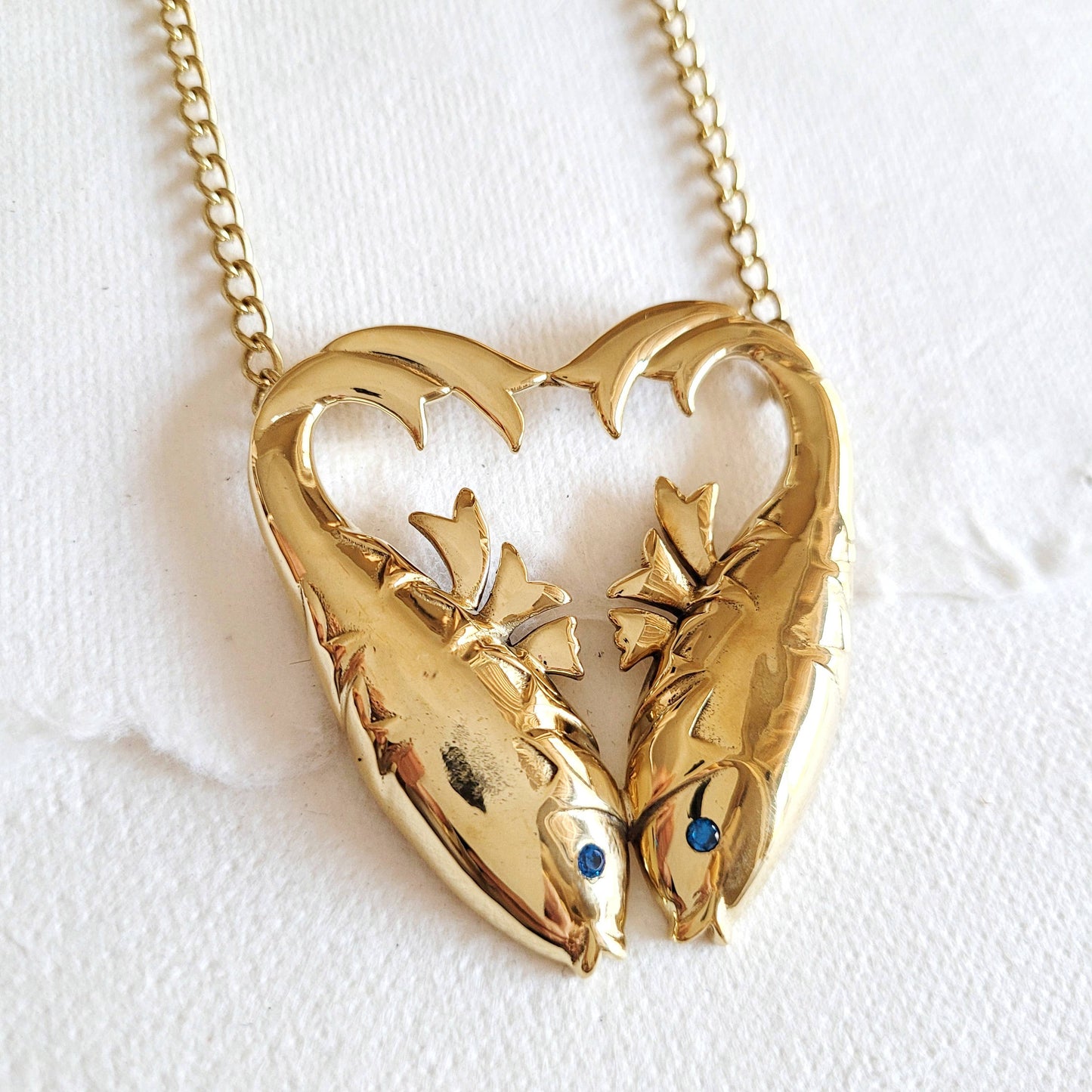 gold double fish heart shaped necklace with inlaid aquamarine stones for the eyes against a white background