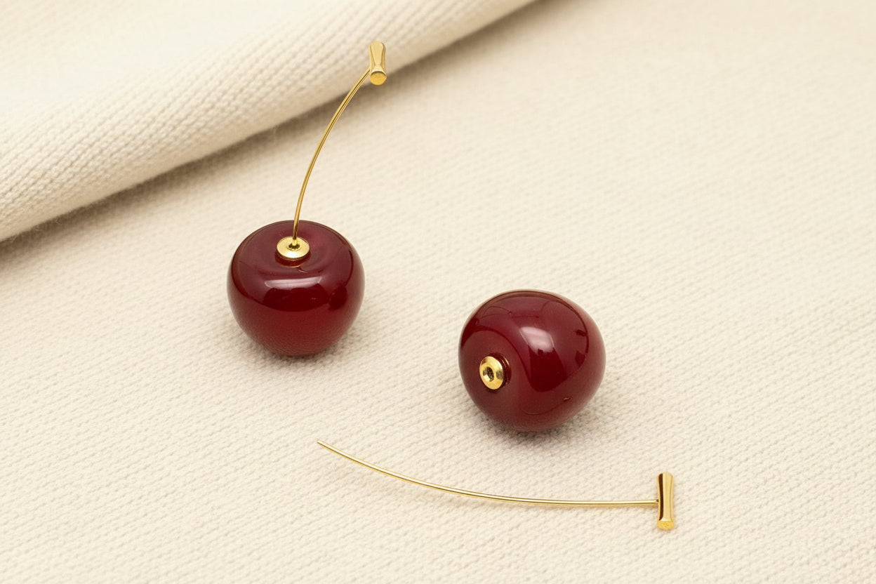 a pair of red cherry resin earrings with gold stems laying on a grey background