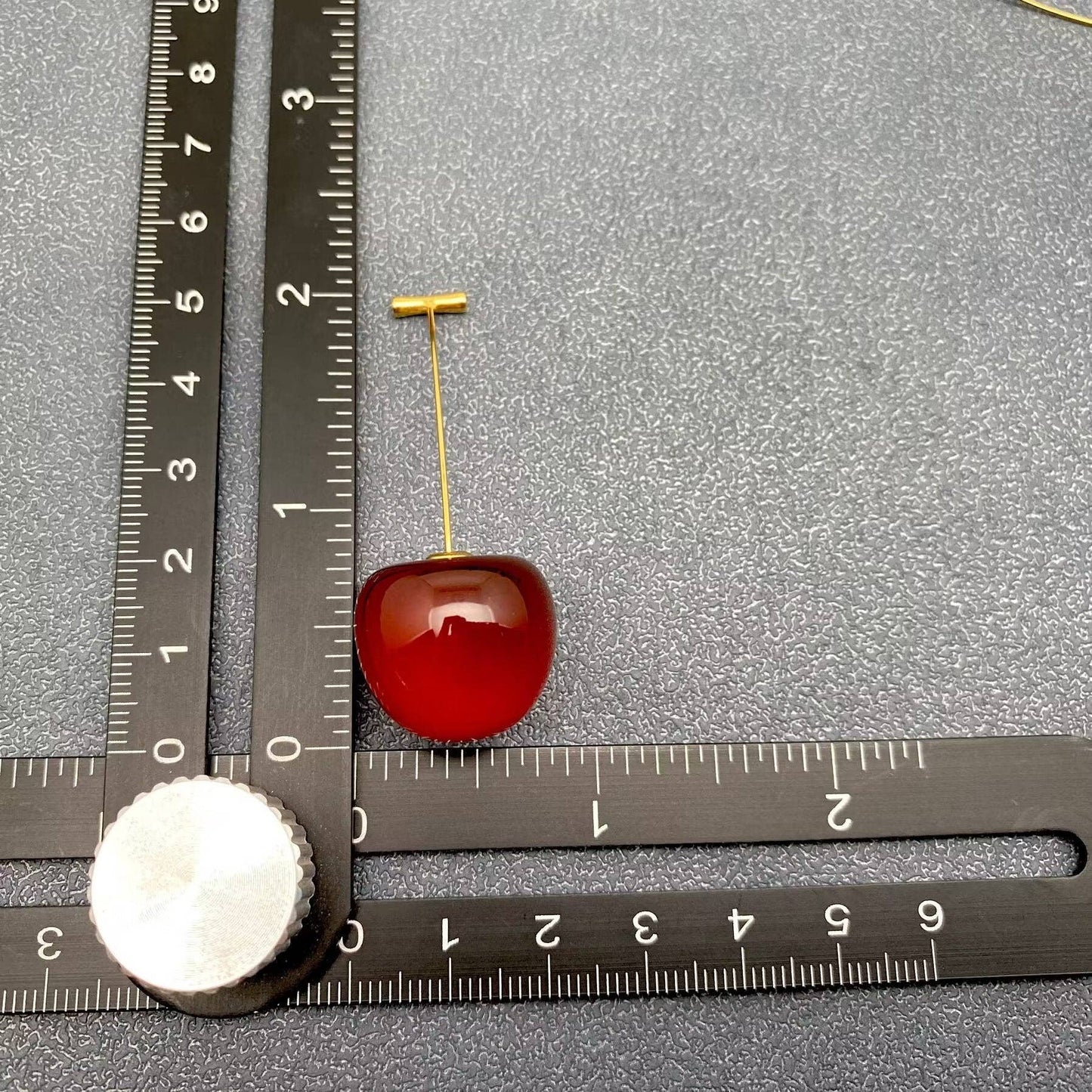 one red cherry themed earring with gold stem next to a black ruler laying on a grey background