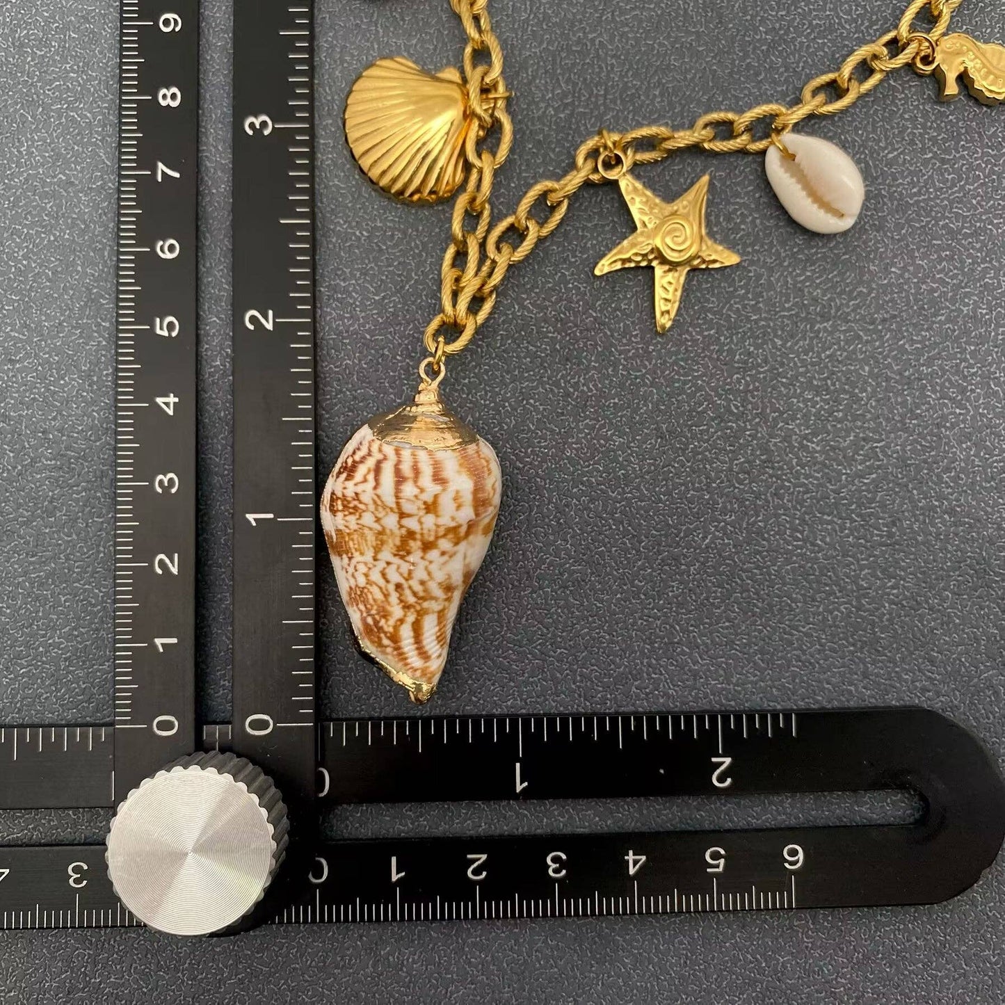 coast themed multi charm necklace with conch shell next to black ruler to show the length of the conch shell