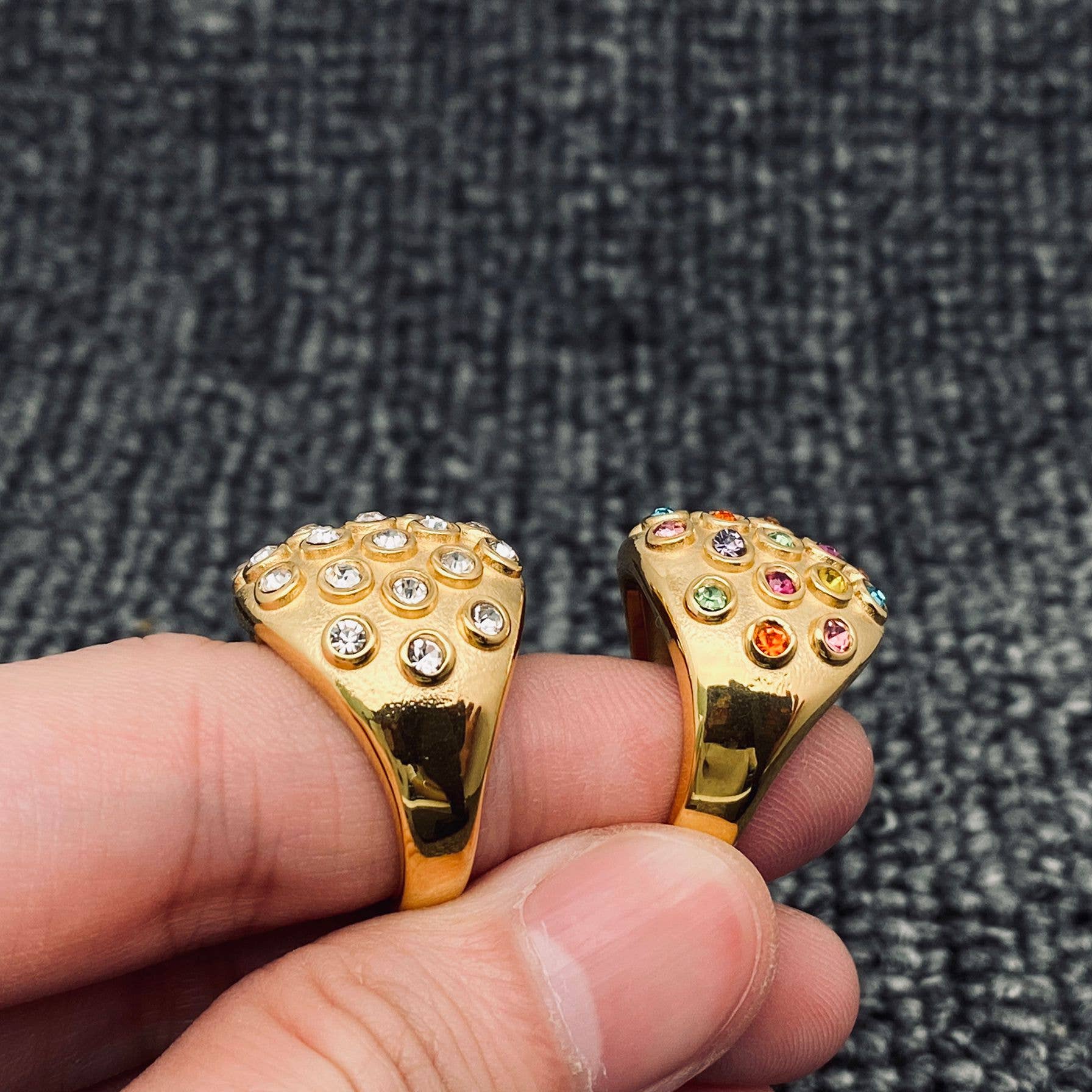Fingers showing the side view of 2 gold statement rings. One ring has clear crystal stones and the other ring has multi colored crystal stones.