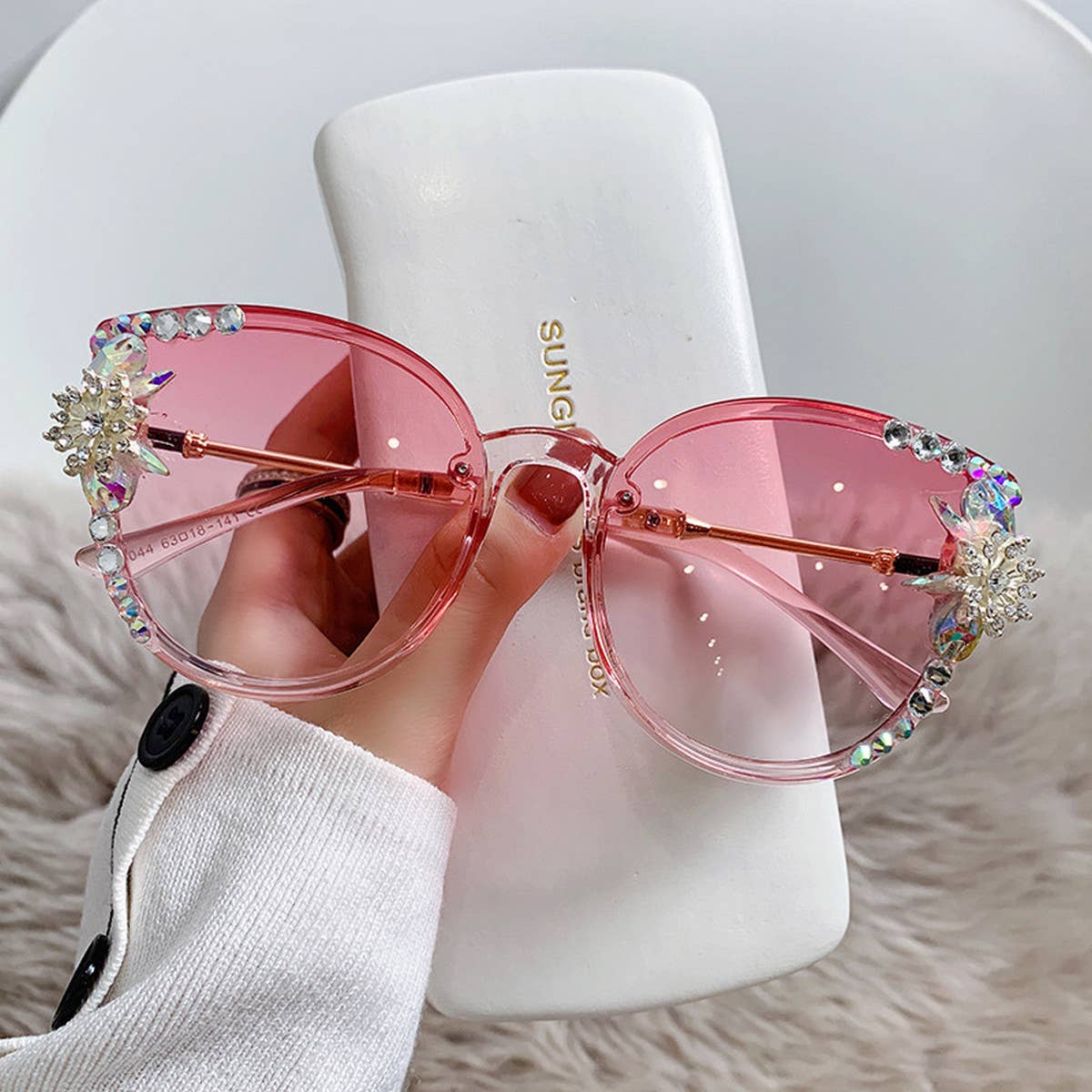 a woman's hand holding crystal embellished cat eye sunglasses with pink lens and a white sunglass case against an ivory faux fur background