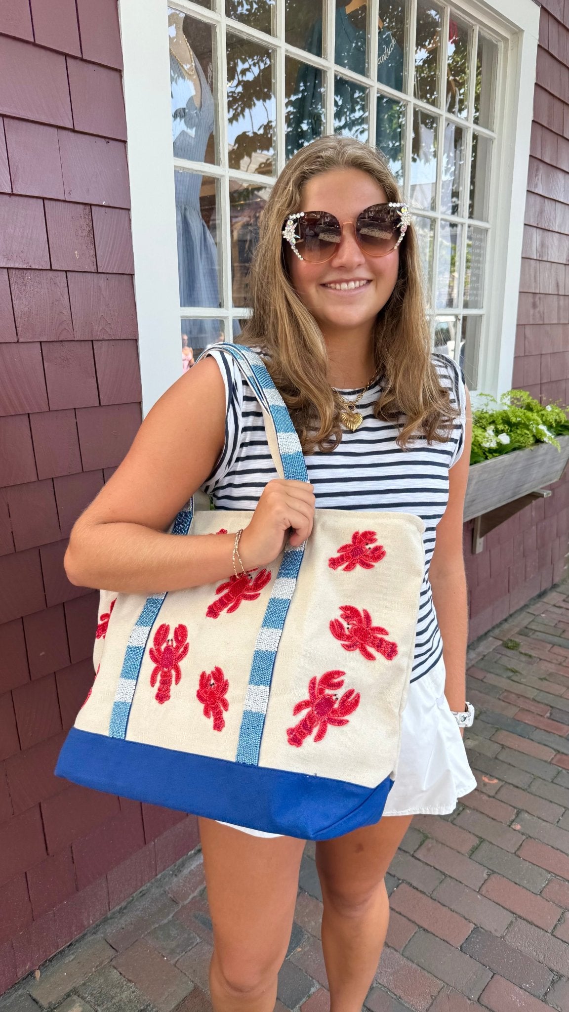 a woman with brown hair wearing crystal embellished cat eye sunglasses with a blue and white striped top and lobster beaded tote