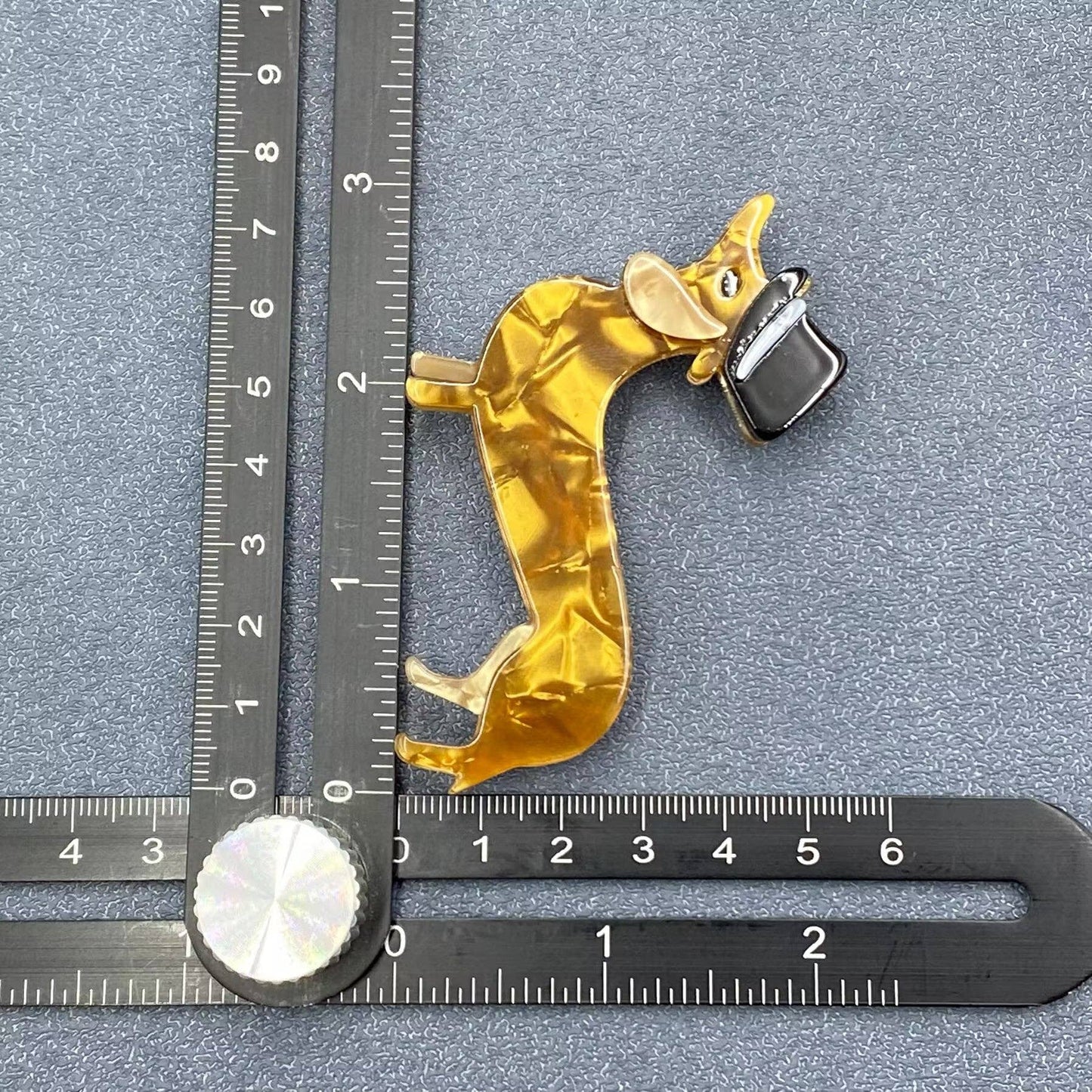 Gold-colored acrylic dog brooch with a black top hat on a gray background with a ruler for scale.
