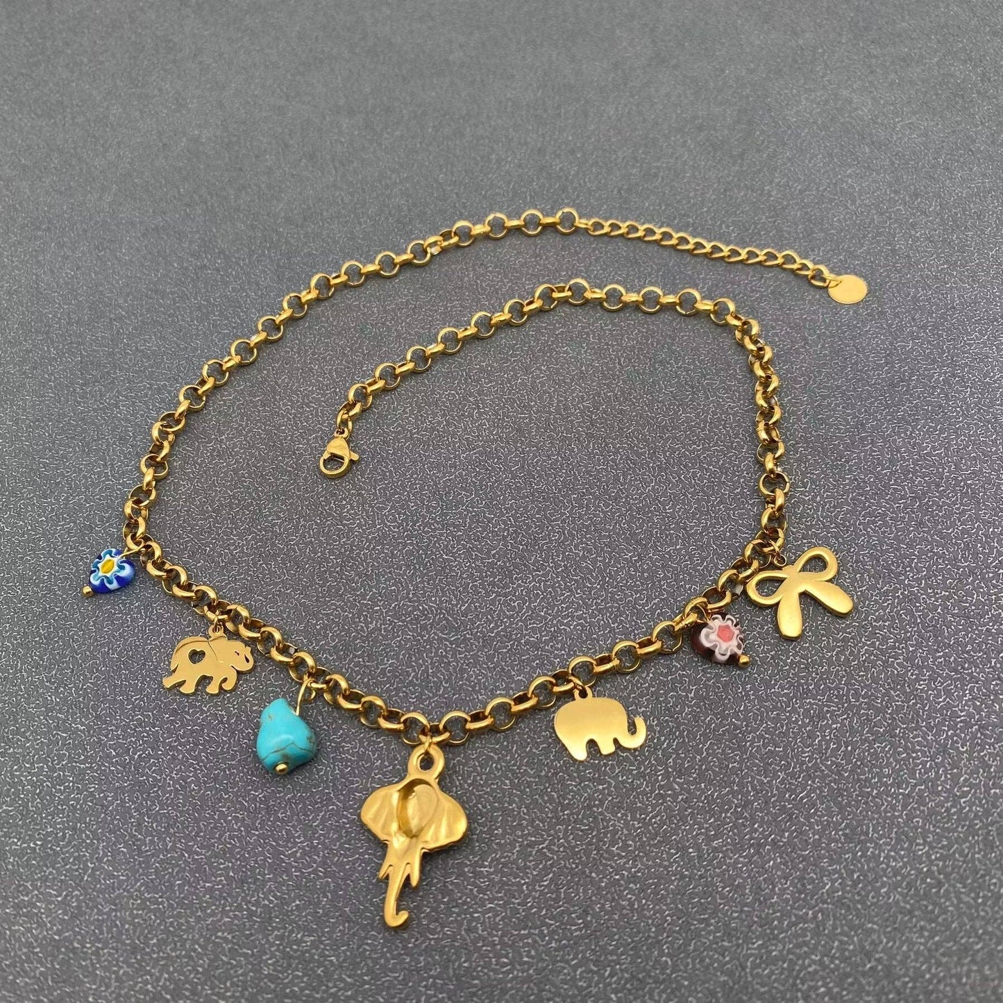 back view of gold multi charm necklace with elephants, bow, and turquoise charms laying on a grey background