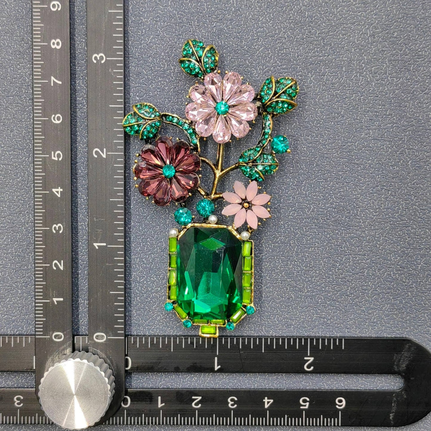 Decorative brooch with flowers and green gemstone next to a ruler for scale on a gray background