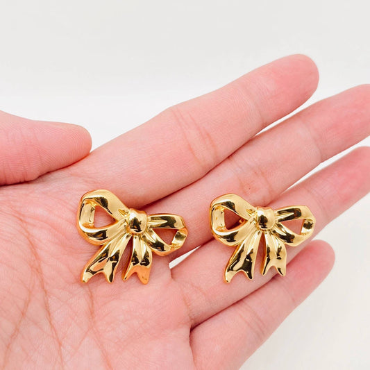 a hand holding a pair of gold bow stud earrings