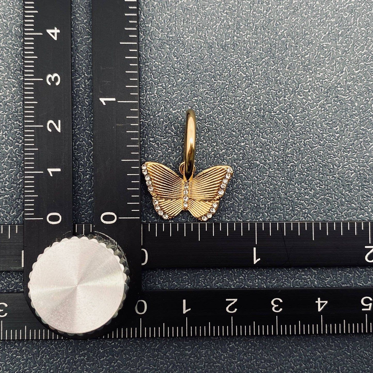 One gold butterfly crystal earring on a textured surface with a ruler for scale.