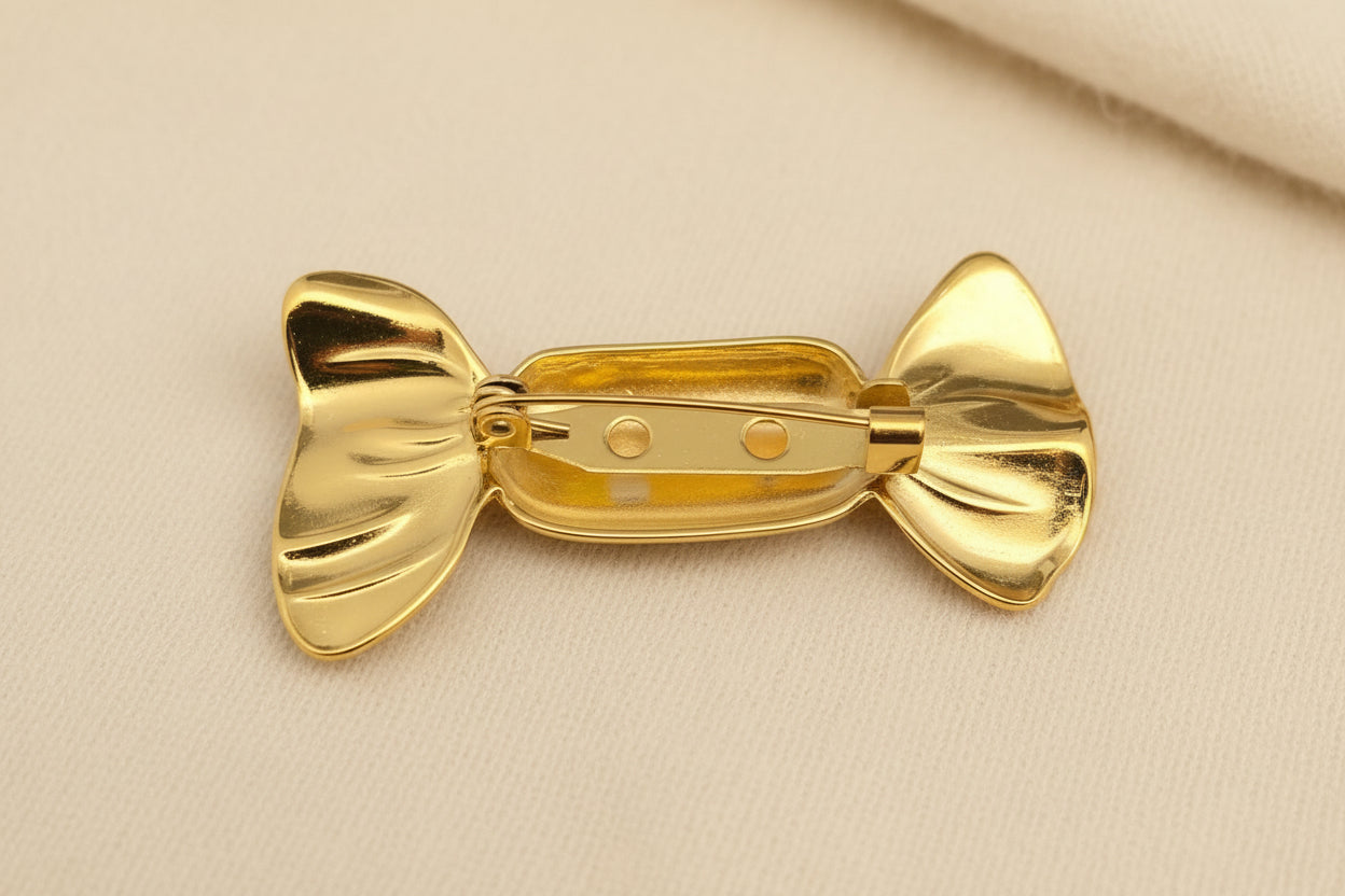Back view of gold candy wrapper-shaped brooch showing pin closure on a gray textured background