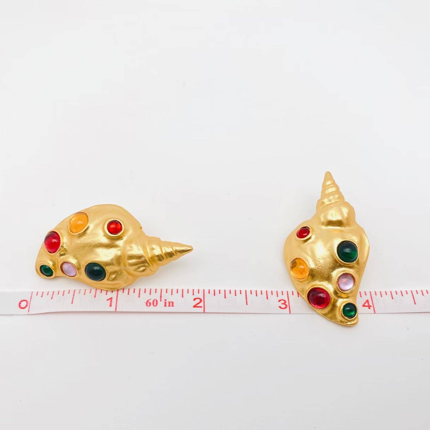 a pair of gold conch earrings with inlaid multi-colored glass stones against a ruler