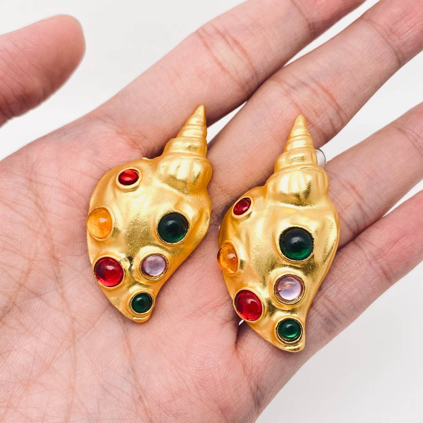 gold conch shaped earrings with inlaid multi glass stones on human hand