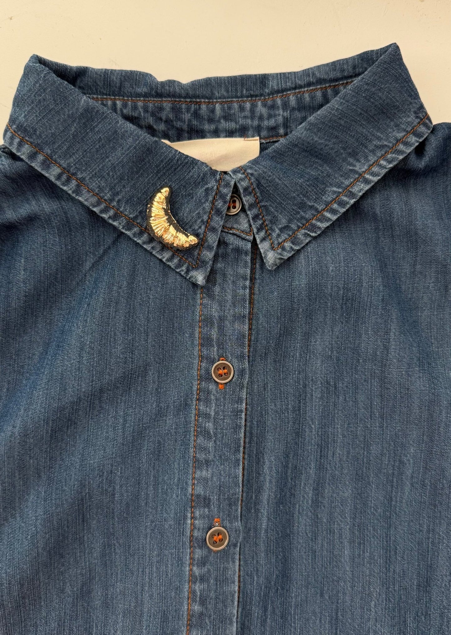 Close-up of a blue denim shirt with a gold croissant brooch on the collar.