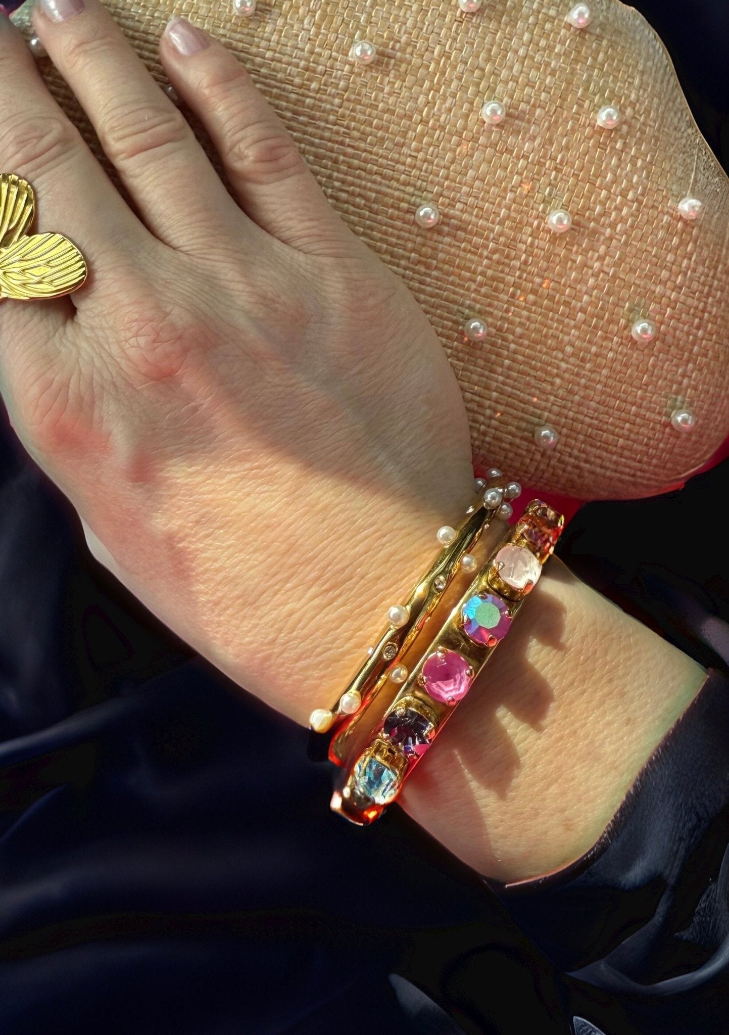 Hand wearing a gold bracelet with colorful stones and a gold ring on a black fabric background.