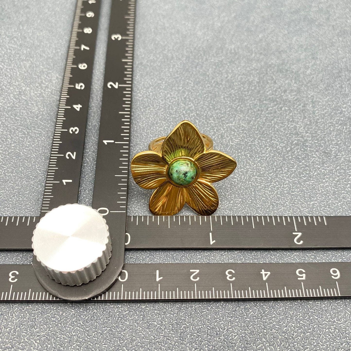 gold flower shaped cocktail ring with green center stone next to a ruler on gray surface