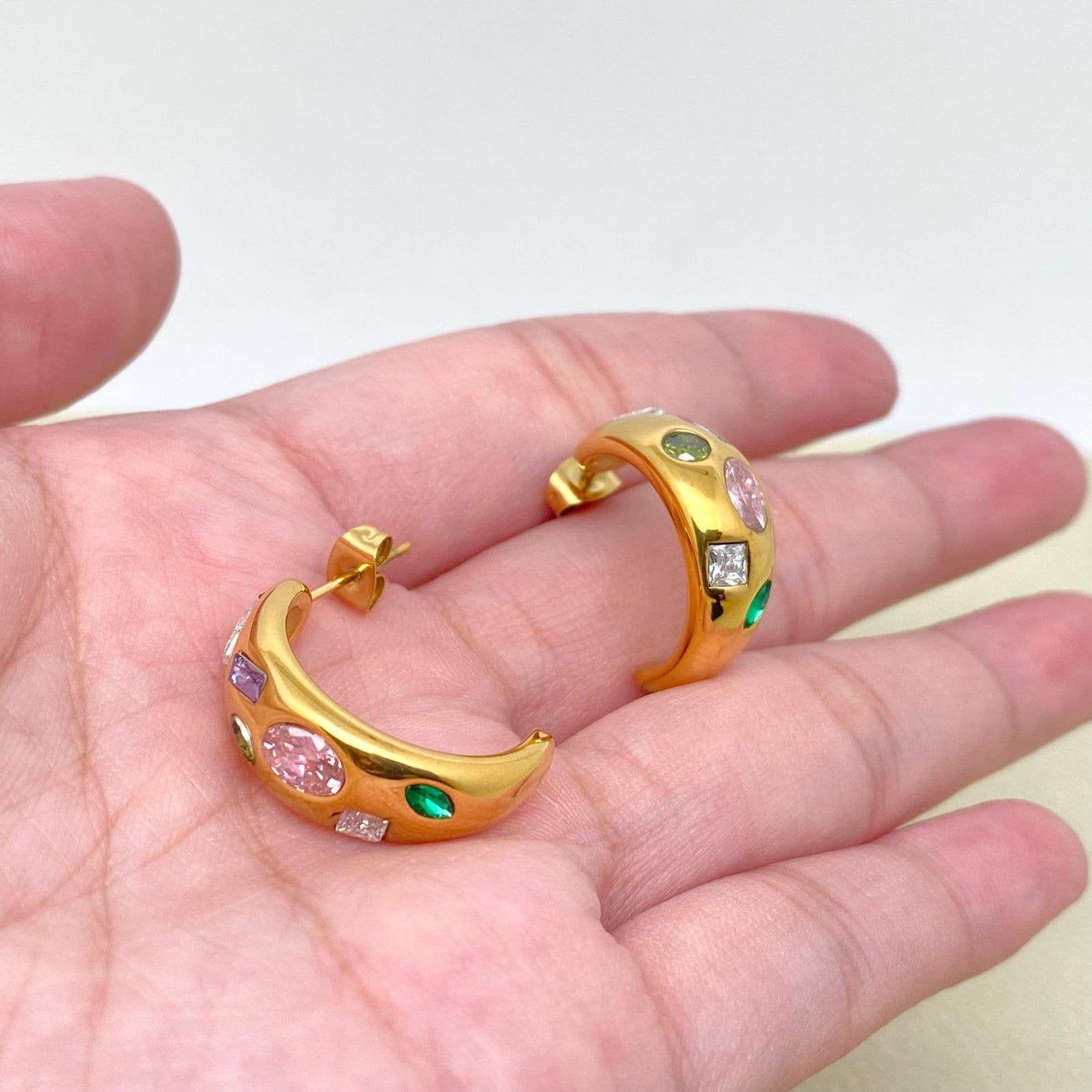 a hand holding a pair of gold multi color crystal inlaid hoop earrings
