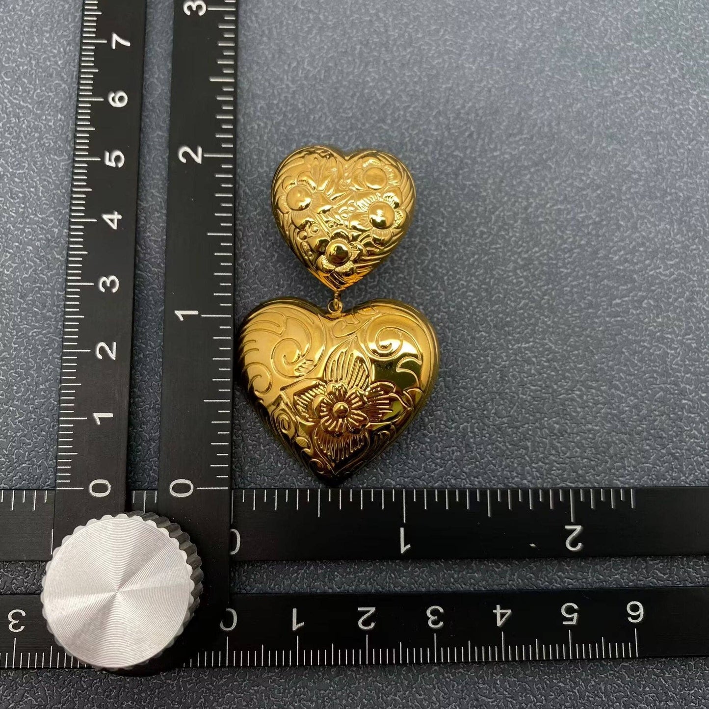 one gold embossed double heart earring next to a black ruler against a grey background