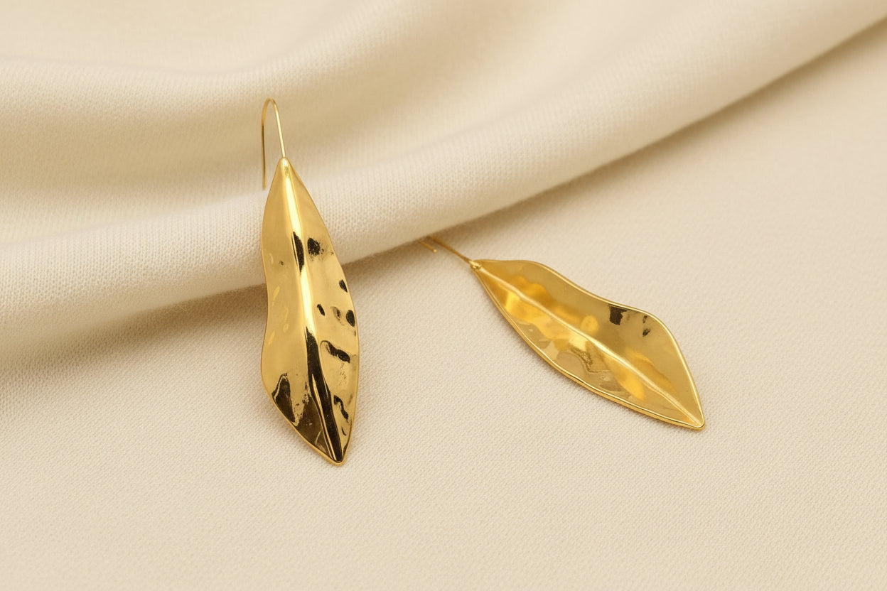 the front and back view of a pair of gold hammered leaf dangle earrings laying on an ivory fabric background