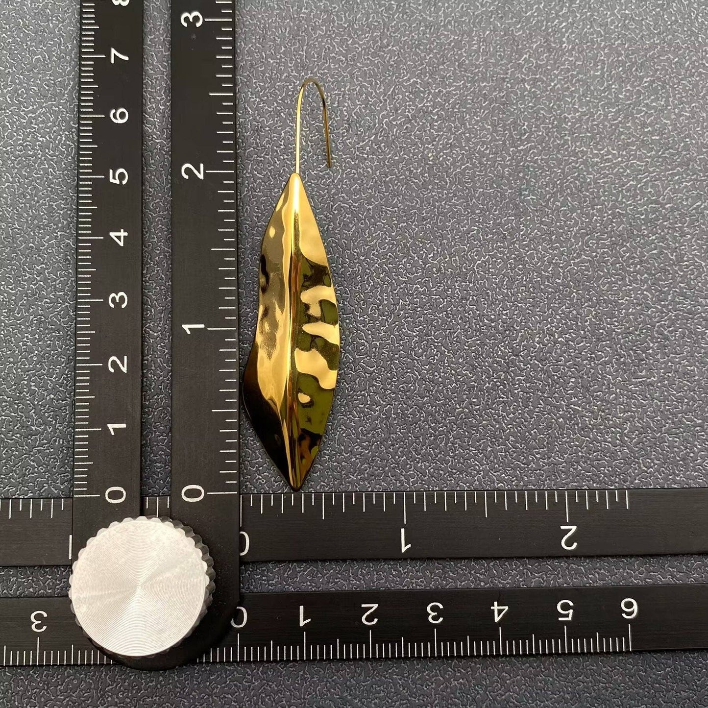 one gold hammered leaf dangle earring next to a black ruler laying on a grey background