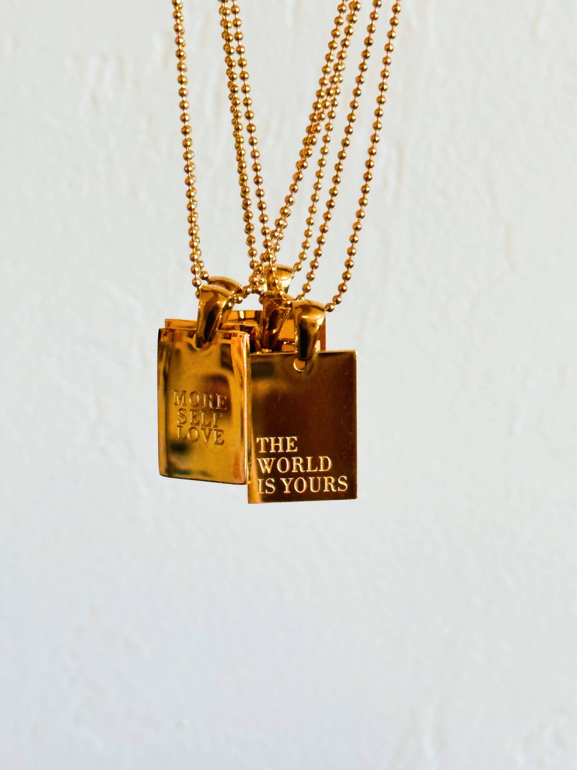gold necklaces with rectangular pendants with "The World is Yours" engraved on one and "More Self Love" engraved on another