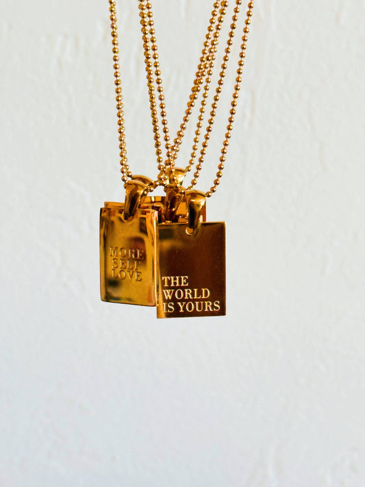 gold necklaces with rectangular pendants with "The World is Yours" engraved on one and "More Self Love" engraved on another