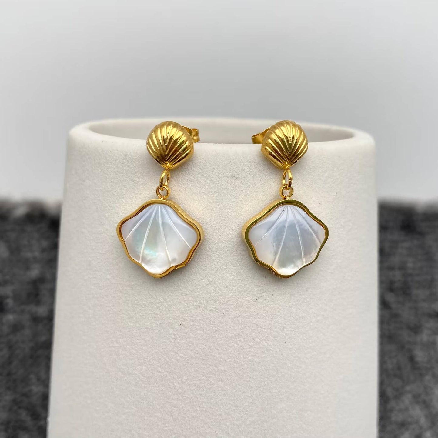 a pair of gold shell charm drop earrings with white resin etched shell design hanging on a white vase