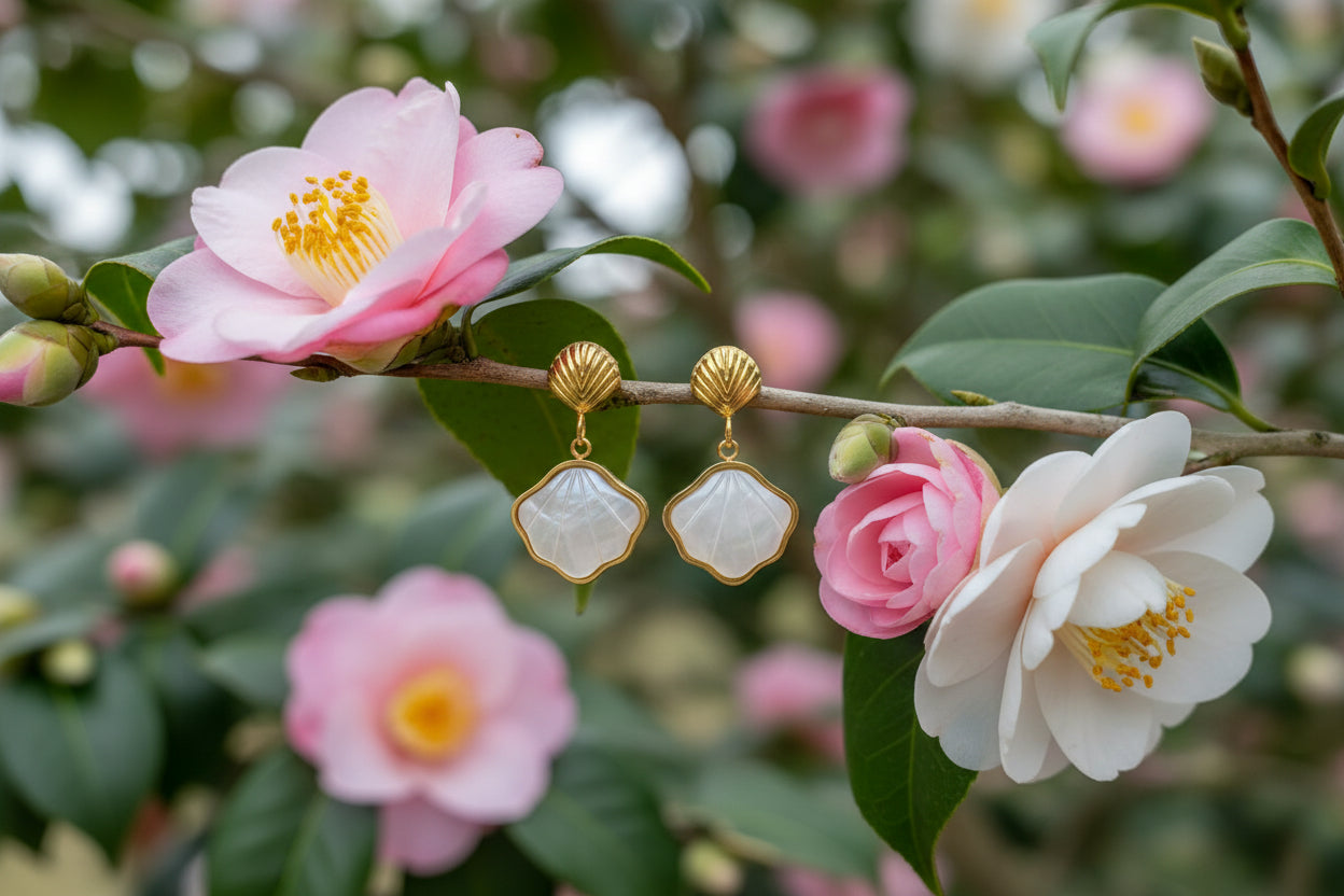 a pair of gold shell charm drop earrings with white resin etched shell design hanging on a camellia flower branch