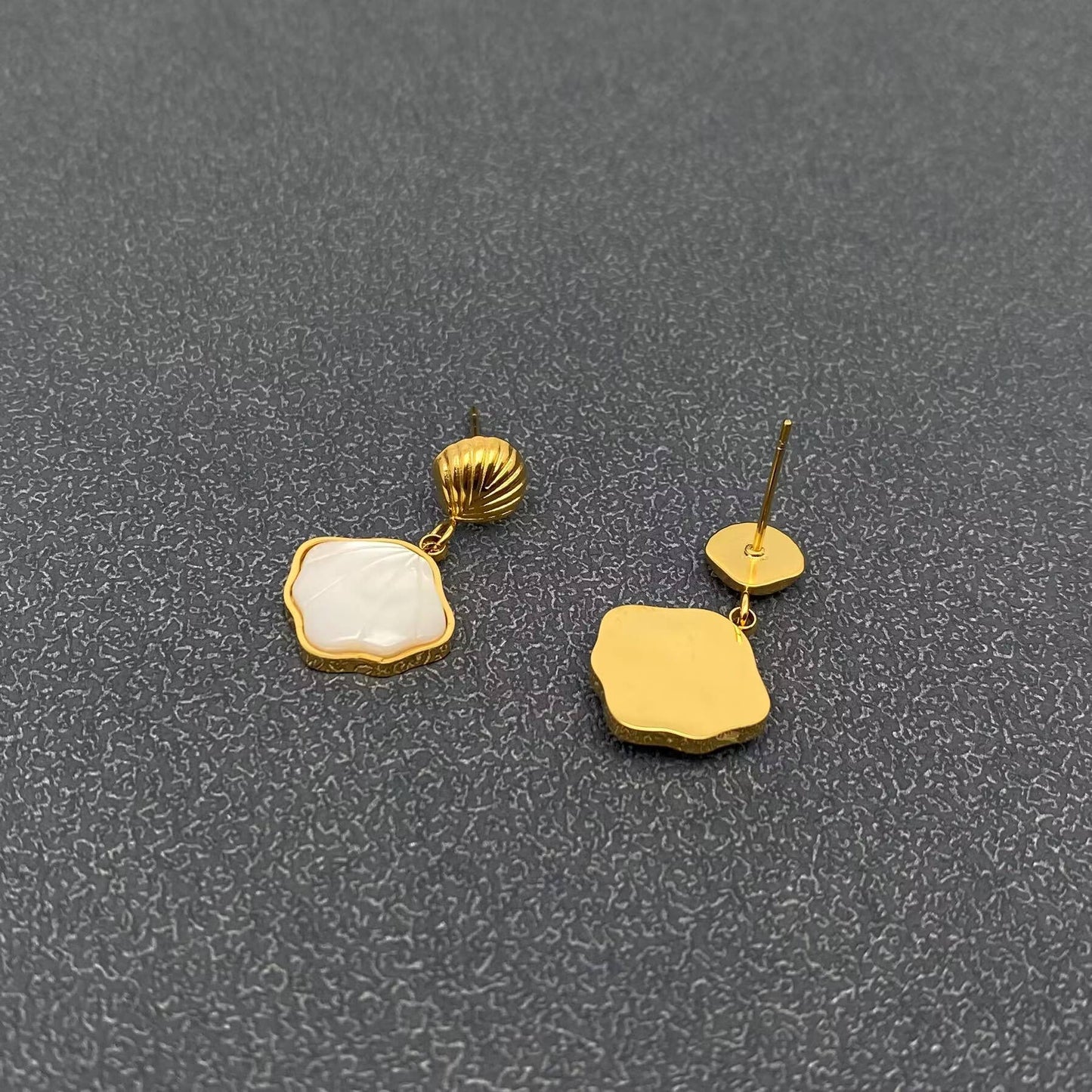the front and back view of a pair of gold shell charm drop earrings with white resin etched shell design laying on a grey background