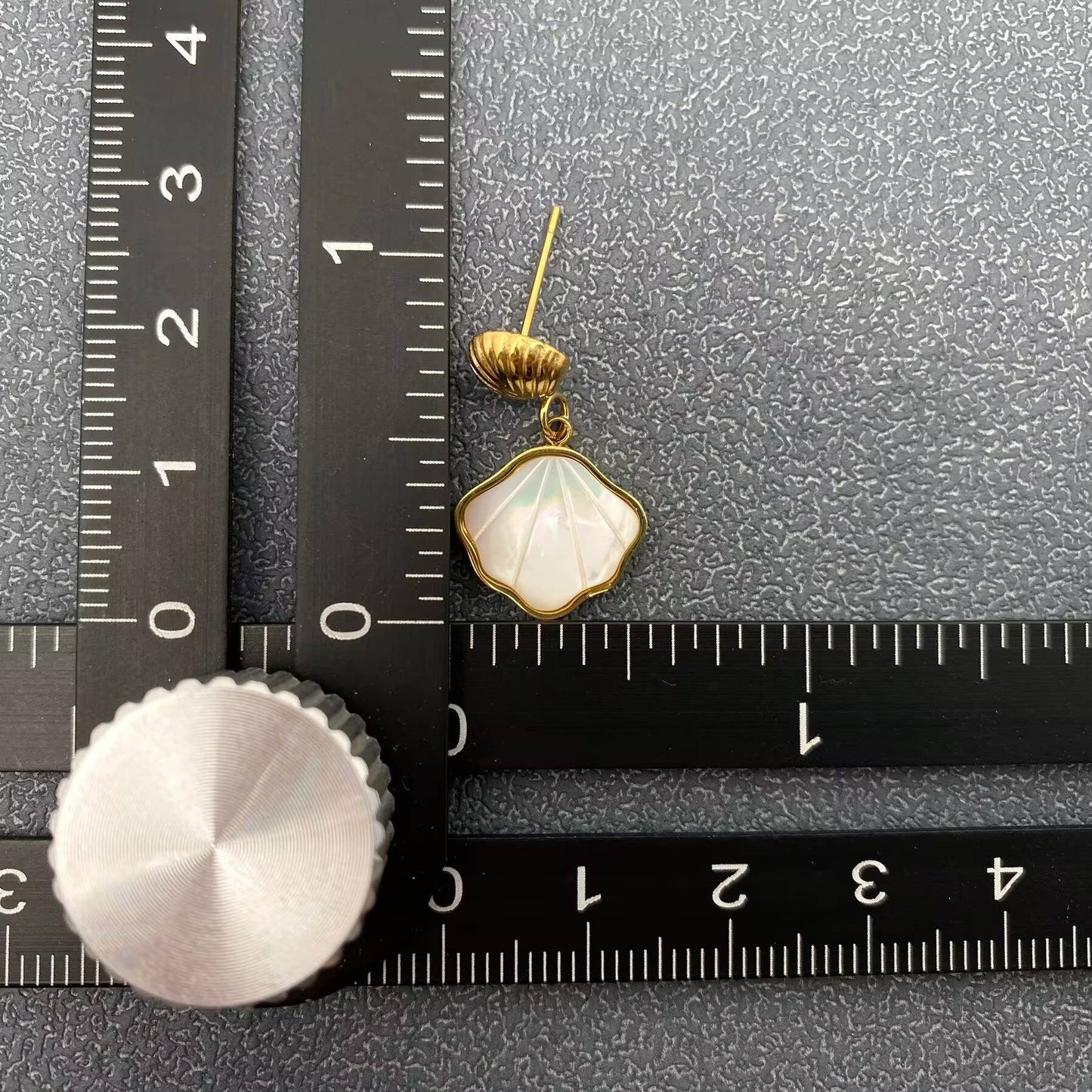 one gold shell charm drop earrings with white resin etched shell design laying next to a black ruler and grey background