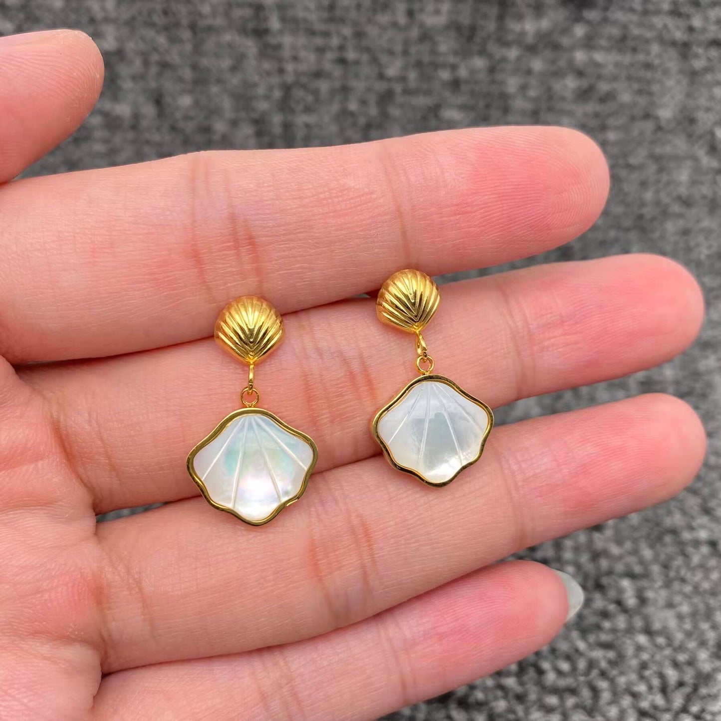 a hand holding a pair of gold shell charm drop earrings with white resin etched shell design