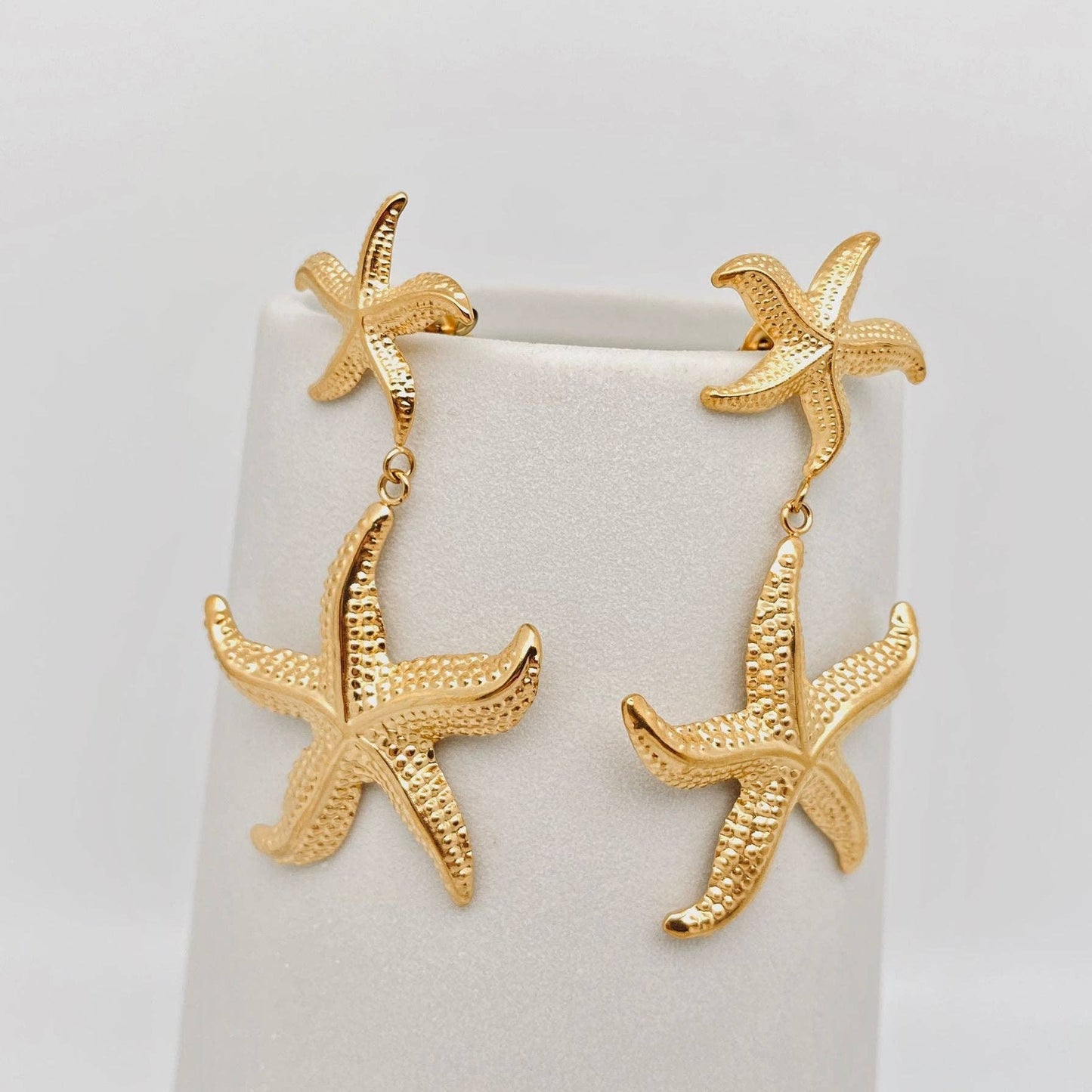 a pair of gold starfish shaped dangle earrings on a white stand