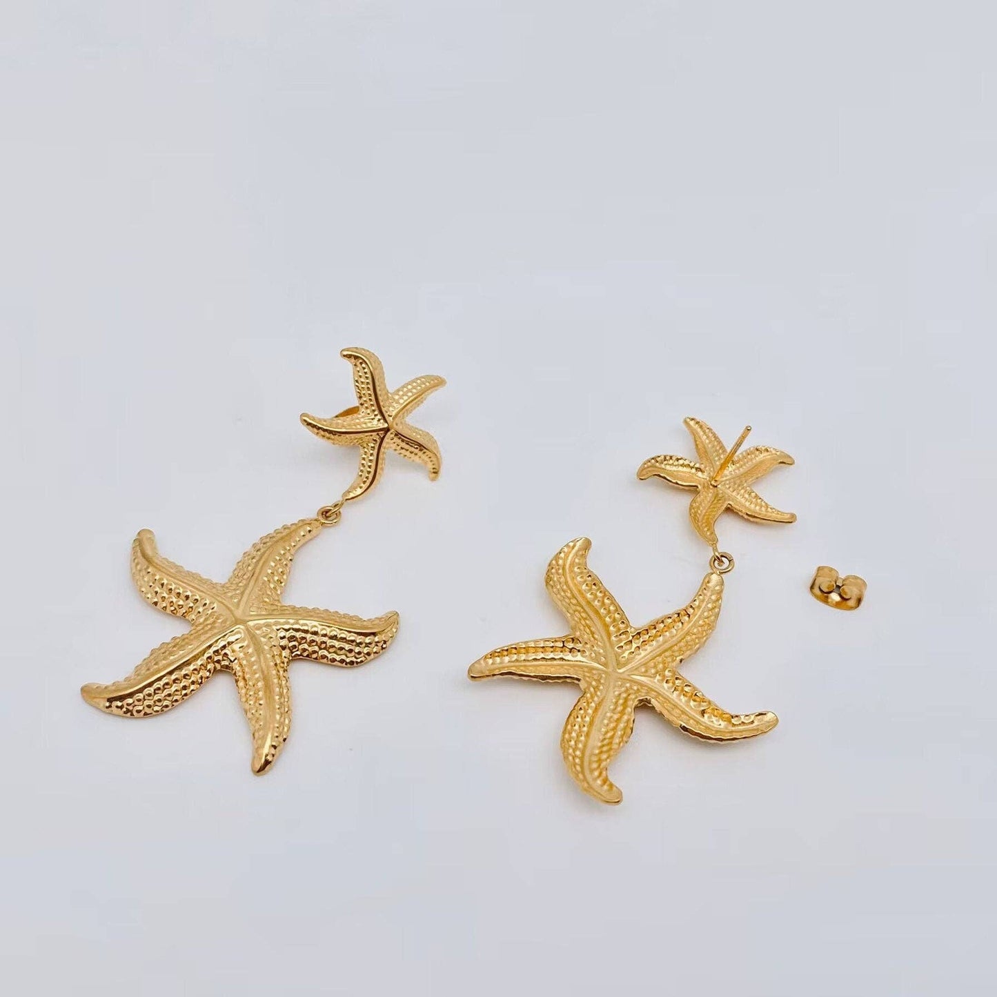 a pair of gold starfish earrings laying on a white background and one shows the front and one shows the back side