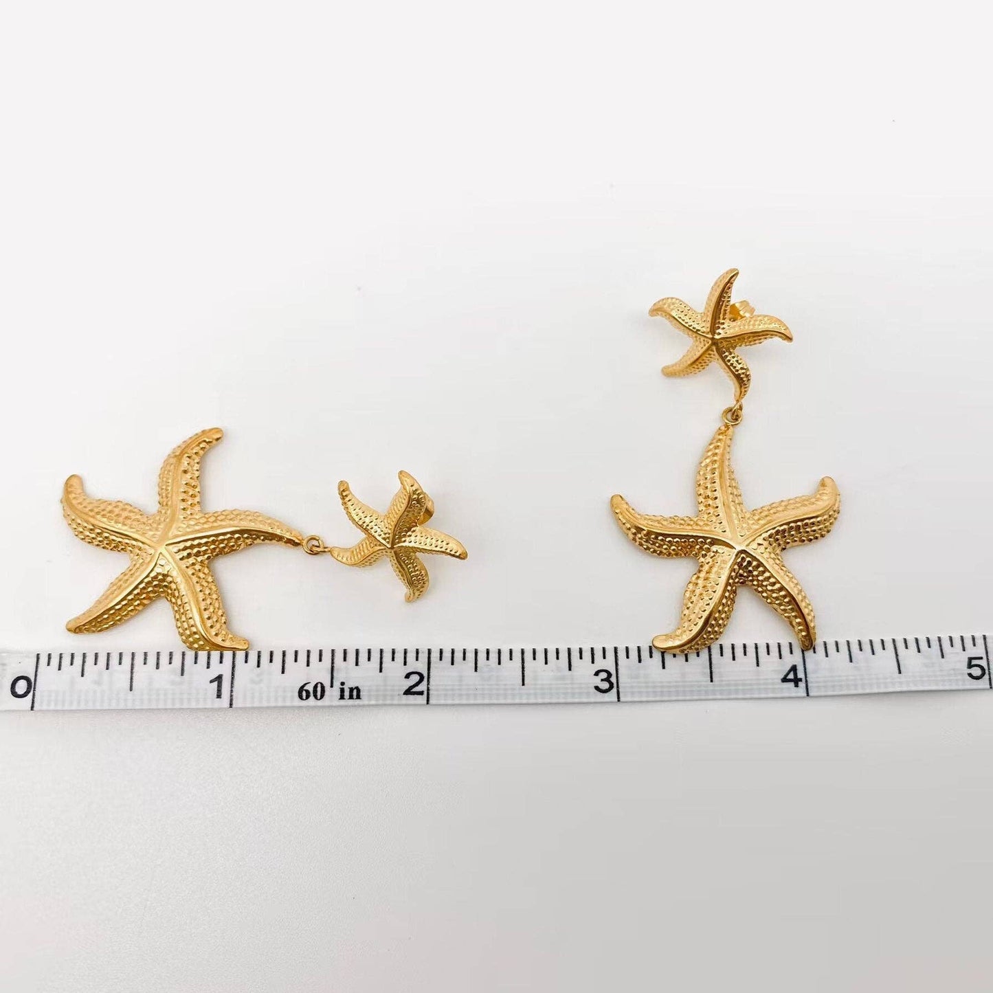a pair of gold starfish shaped earrings laying on a white background next to a ruler to show the length of them
