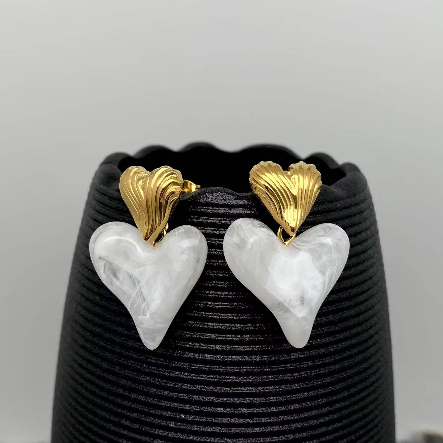 a pair of gold and white resin heart shaped dangle earrings hanging on a black vase