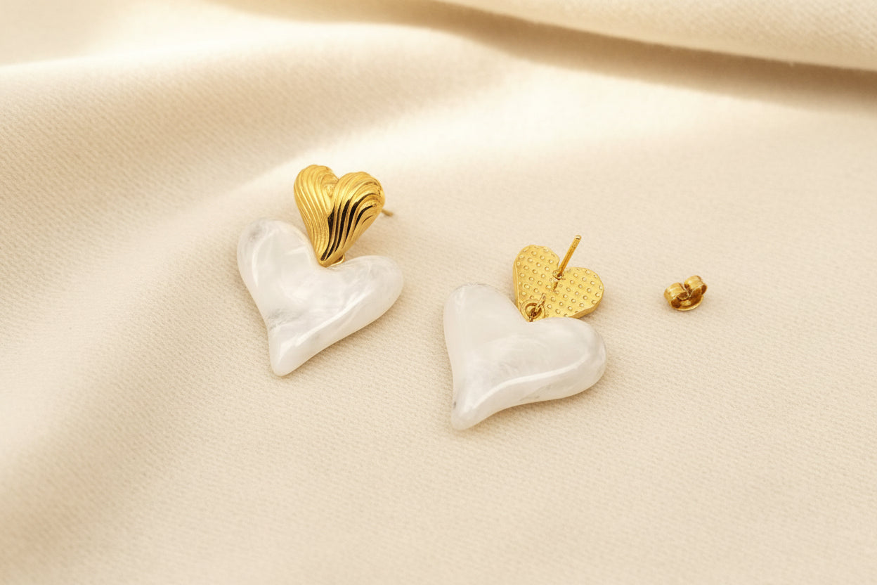 the front and back view of a pair of gold and white resin heart shaped dangle earrings against a grey background