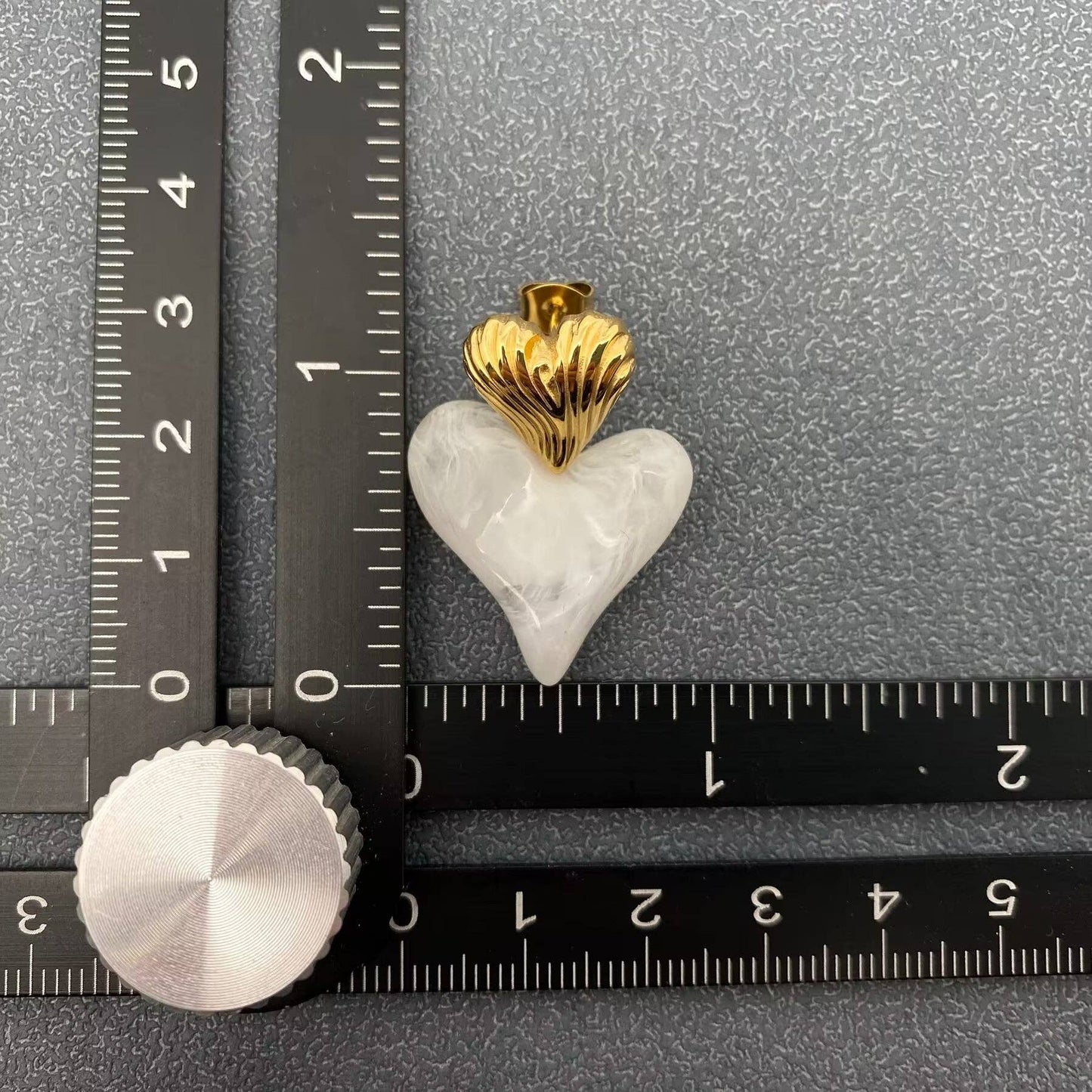 a single gold and white resin heart shaped earring next to a black ruler and a grey background