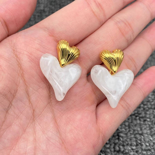 a hand holding a pair of gold and white resin dangle heart earrings