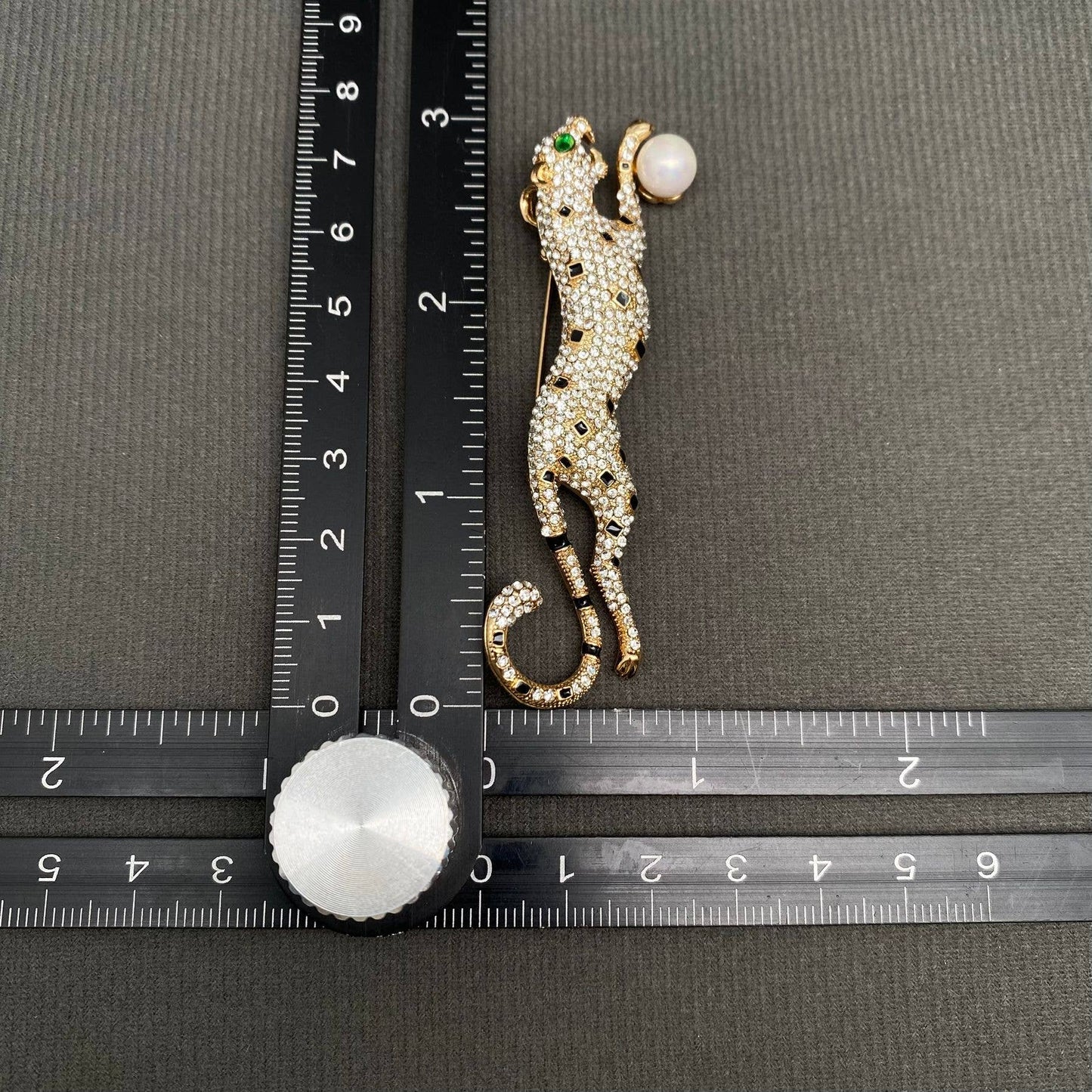 Decorative leopard brooch with gemstones next to a ruler on a gray background