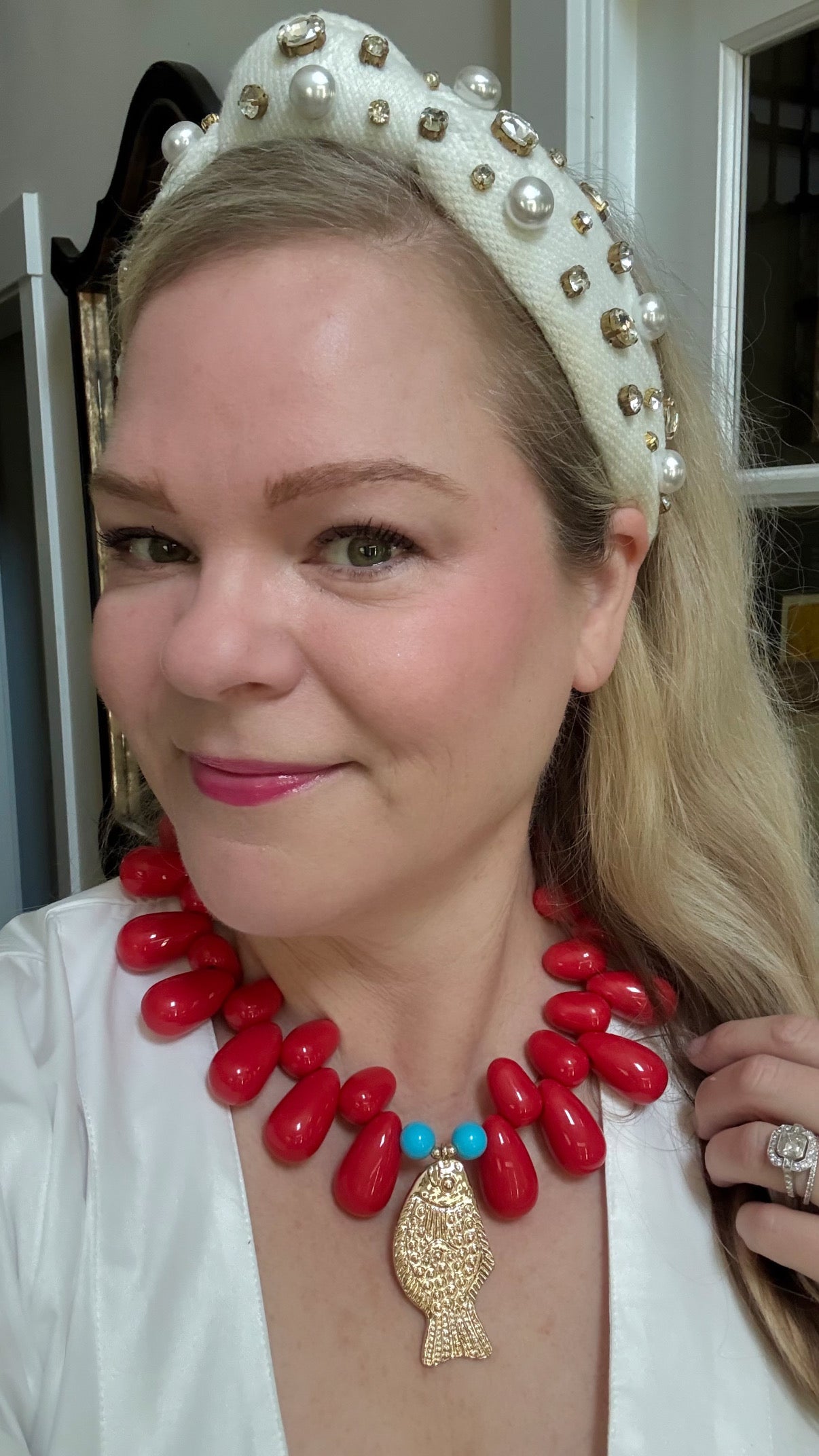 Woman wearing a white headband with pearls, red necklace with a gold fish pendant, and a white top.
