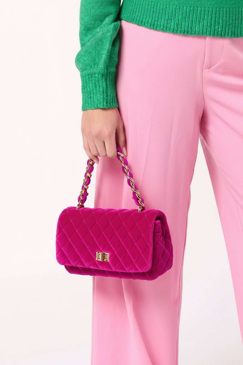 A person wearing pink trousers and a green sweater holds a bright pink quilted handbag with a gold-tone chain strap and a small gold logo plaque on a plain white background.