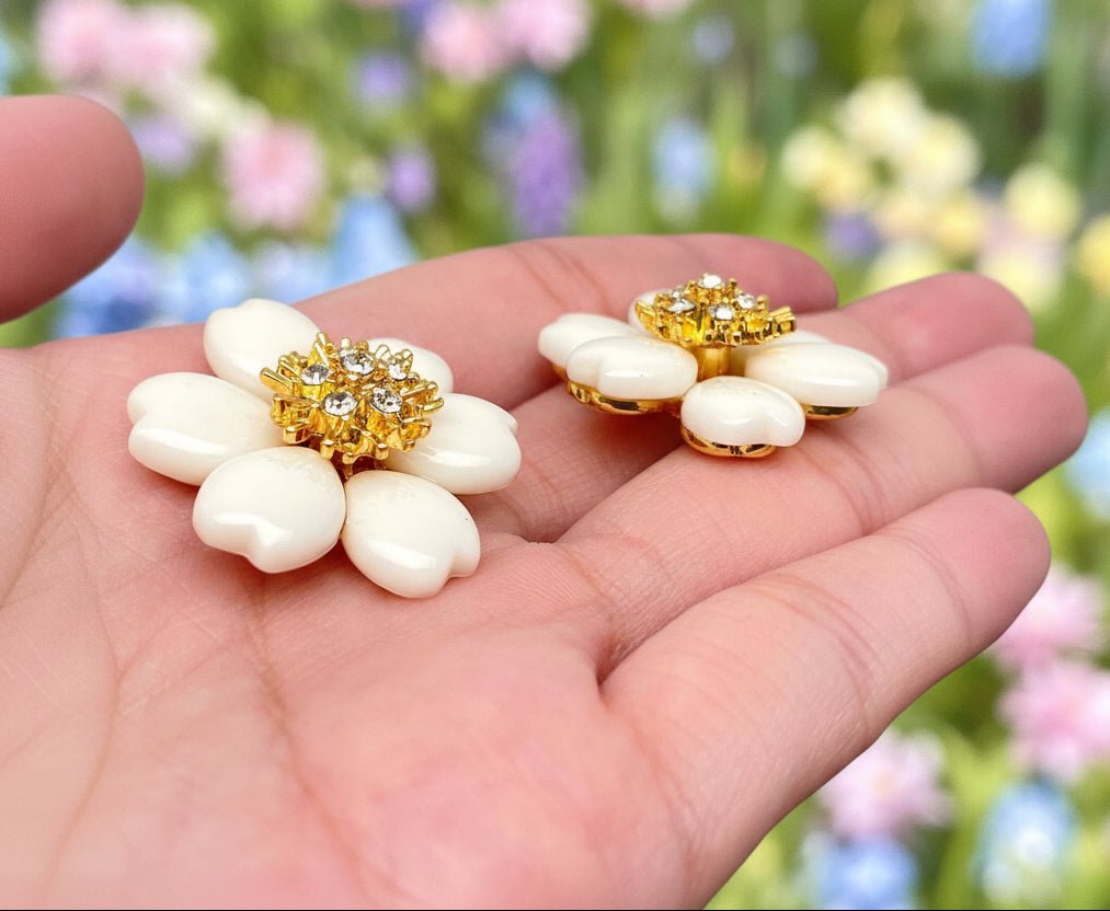 White flower-shaped earrings with gold and crystal details held in a hand and spring garden blurred background