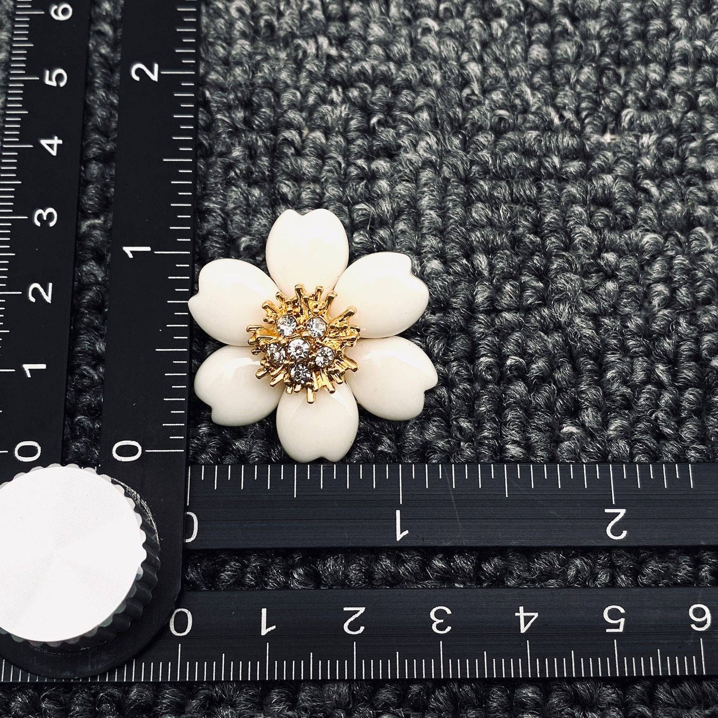 White flower-shaped earrings with gold center on a grey textured surface with ruler for scale.