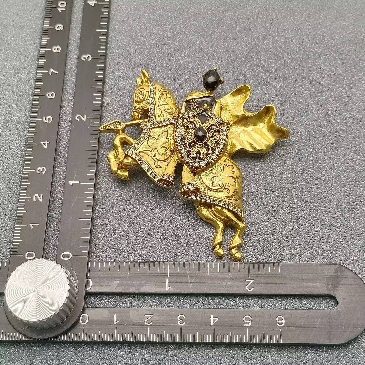 gold and black medieval knight and horse themed brooch next to a ruler for scale against a grey textured background