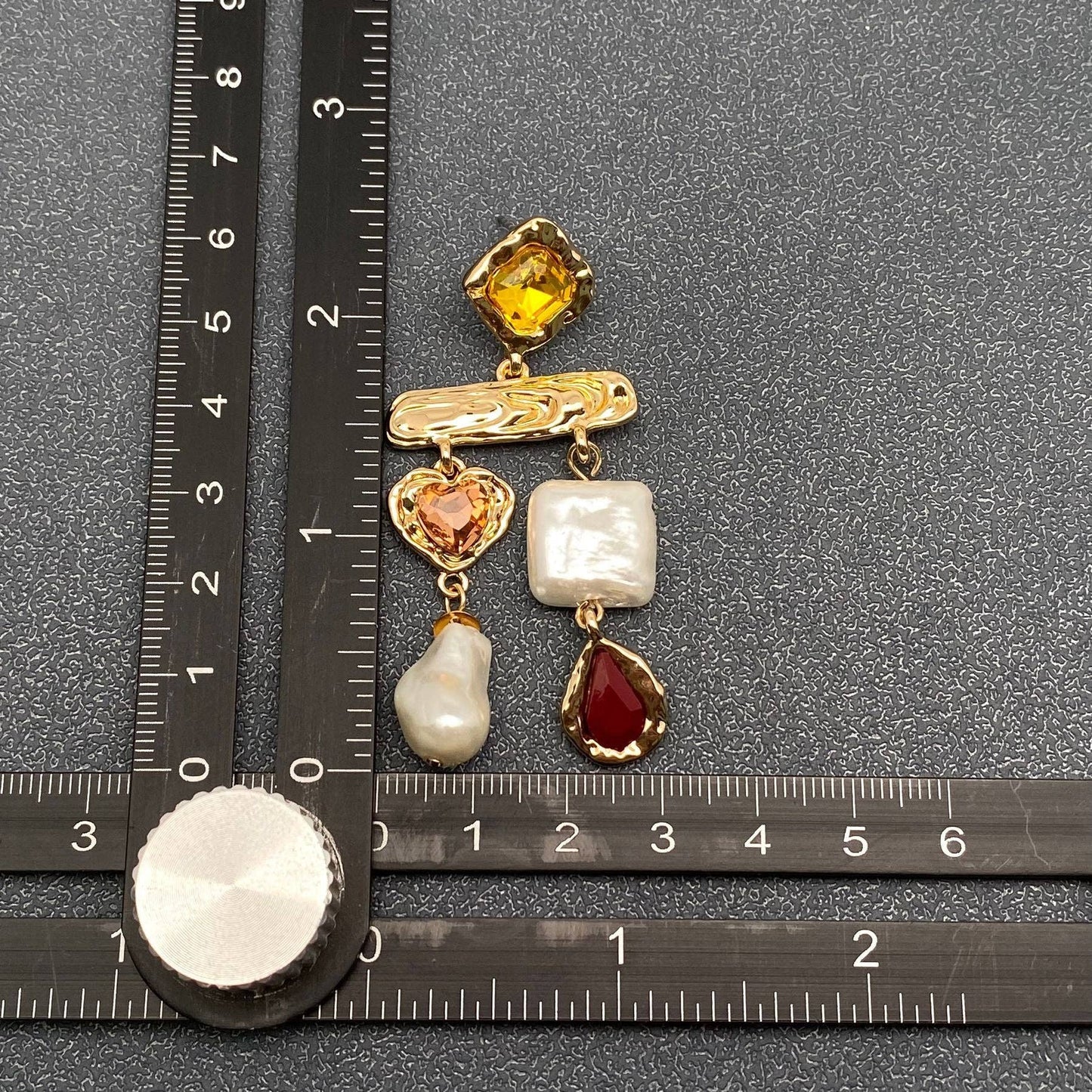 a multi stone baroque pearl earring next to a ruler for scale against a grey background